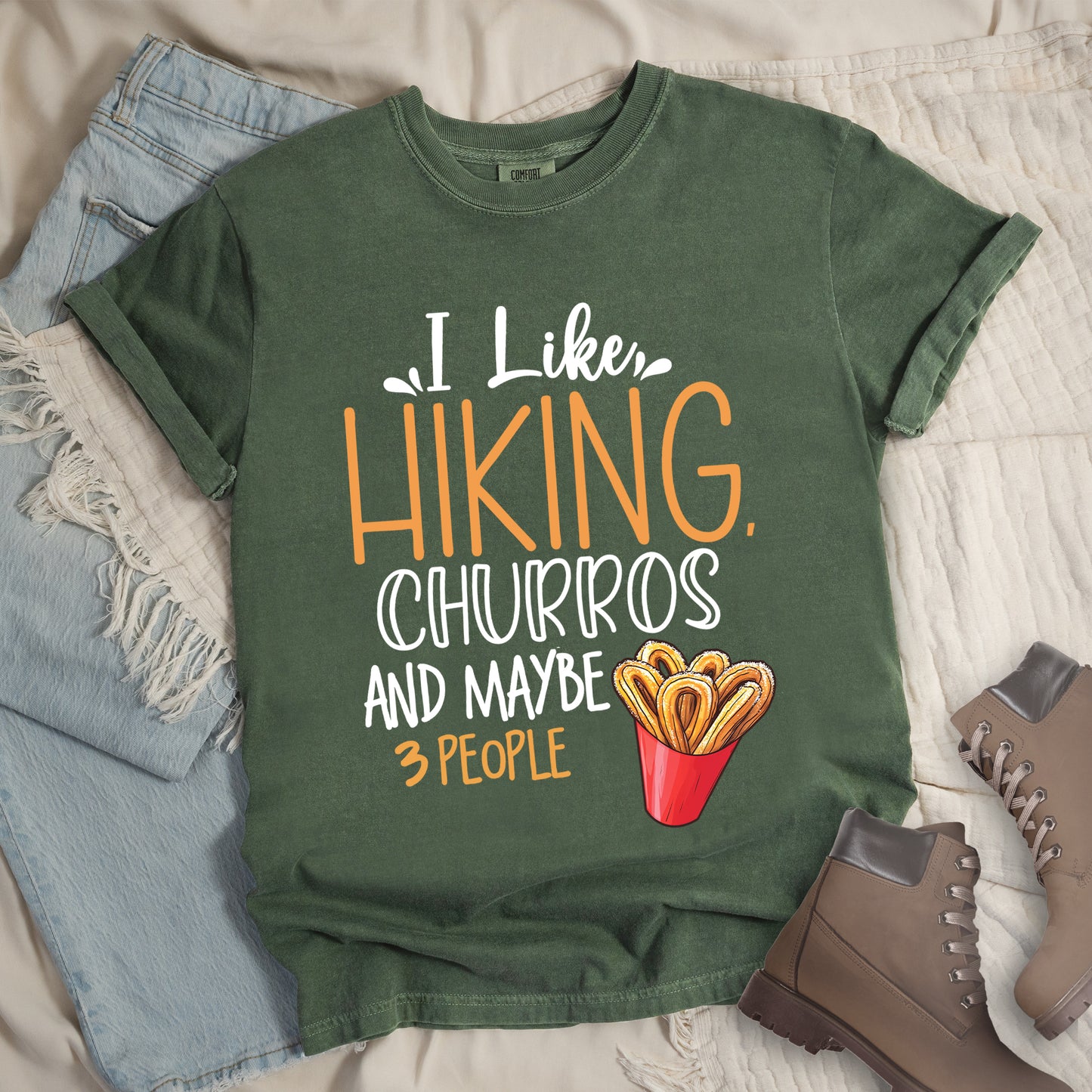 Moss (Dark Green) t-shirt with white and orange text reading "I Like HIKING, CHURROS AND MAYBE 3 PEOPLE". "I Like" appears in white script at the top, "HIKING," in large orange letters, "CHURROS" in white outlined letters, "AND MAYBE" in white, and "3 PEOPLE" in small orange text at the bottom. Features an illustration of churros in a red container on the right side. Features a classic crew neck and casual rolled sleeves, styled alongside light wash denim and brown hiking boots.