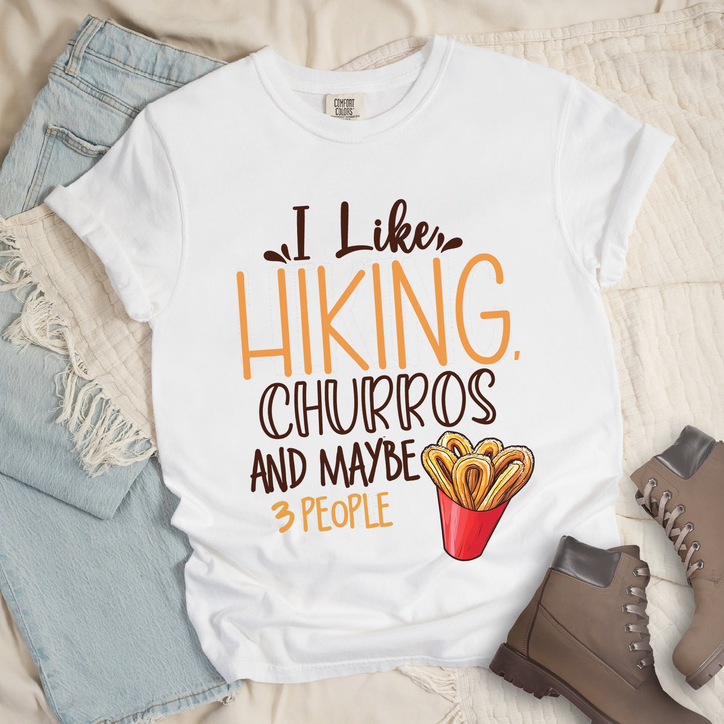 White t-shirt with deep brown and orange text reading "I Like HIKING, CHURROS AND MAYBE 3 PEOPLE". "I Like" appears in deep brown script at the top, "HIKING," in large orange letters, "CHURROS" in deep brown outlined letters, "AND MAYBE" in deep brown, and "3 PEOPLE" in small orange text at the bottom. Features an illustration of churros in a red container on the right side. Features a classic crew neck and casual rolled sleeves, styled alongside light wash denim and brown hiking boots.