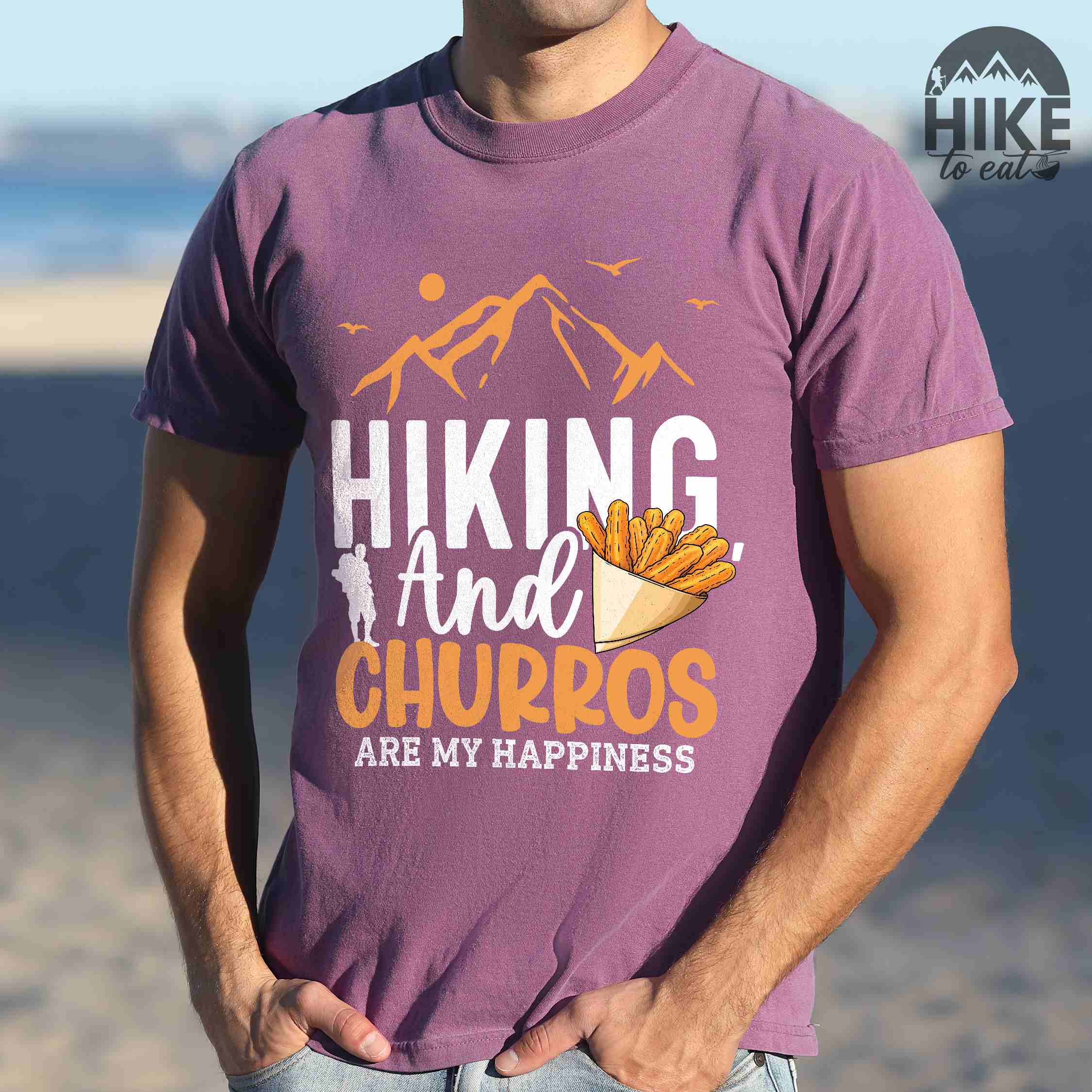 Man wearing berry "hiking and churros are my happiness " colored vintage tee with bold typography, mountains, birds, illustrated a serving of Churros, and backpackers with a sweet tooth.