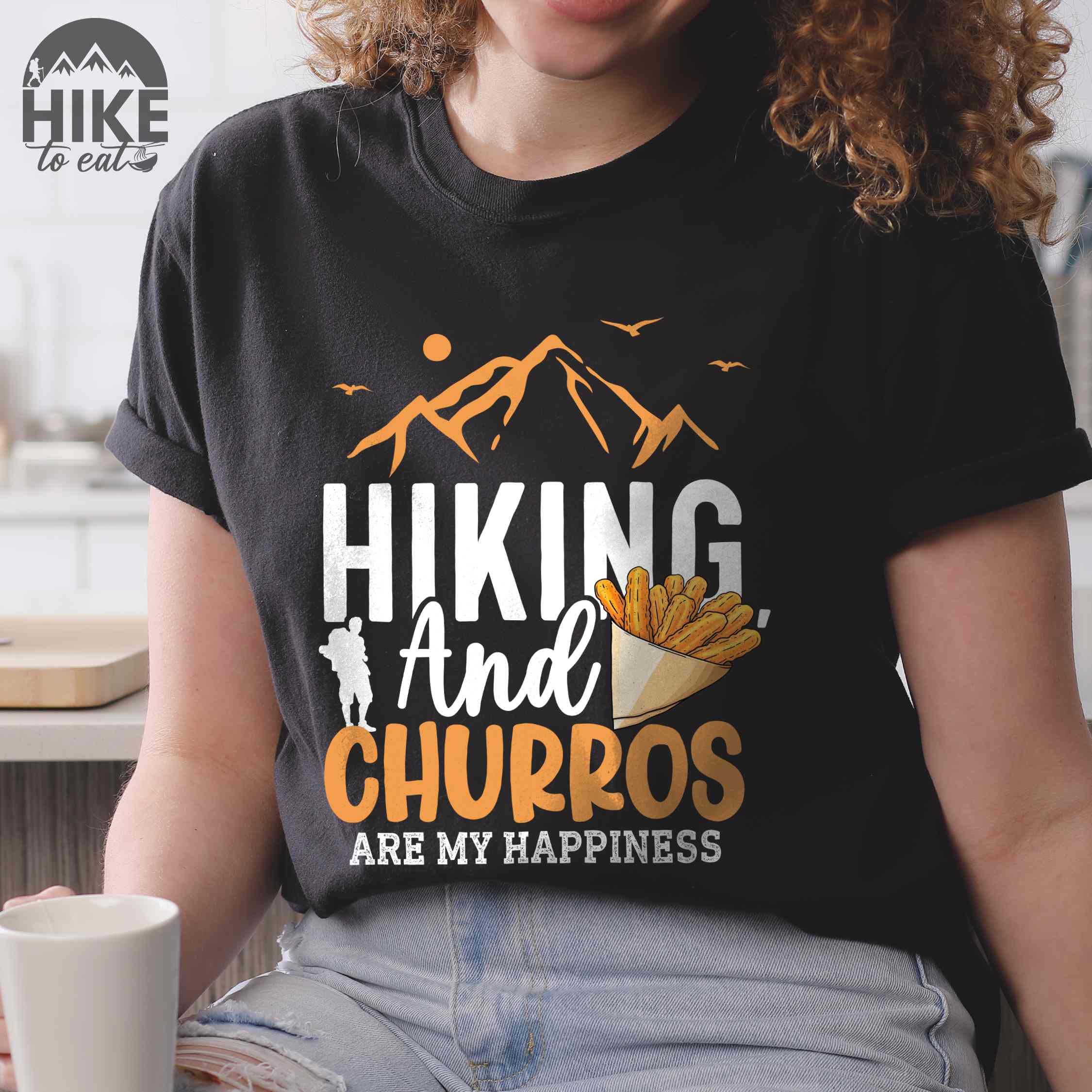 Woman wearing black "hiking and churros are my happiness " colored vintage tee with bold typography, mountains, birds, illustrated a serving of Churros, and backpackers with a sweet tooth.