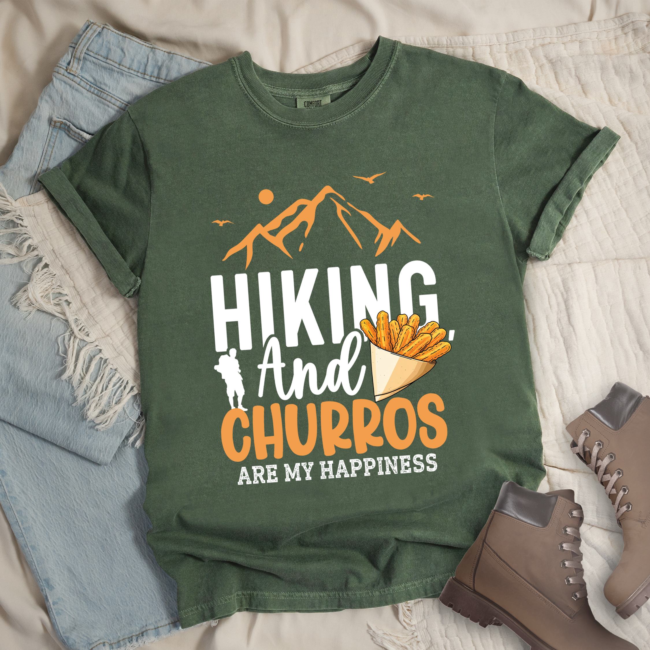 Dark green shirt with the phrase “Hiking And Churros Are My Happiness.” Features a hiker silhouette, mountains, birds, and a churros cone illustration.