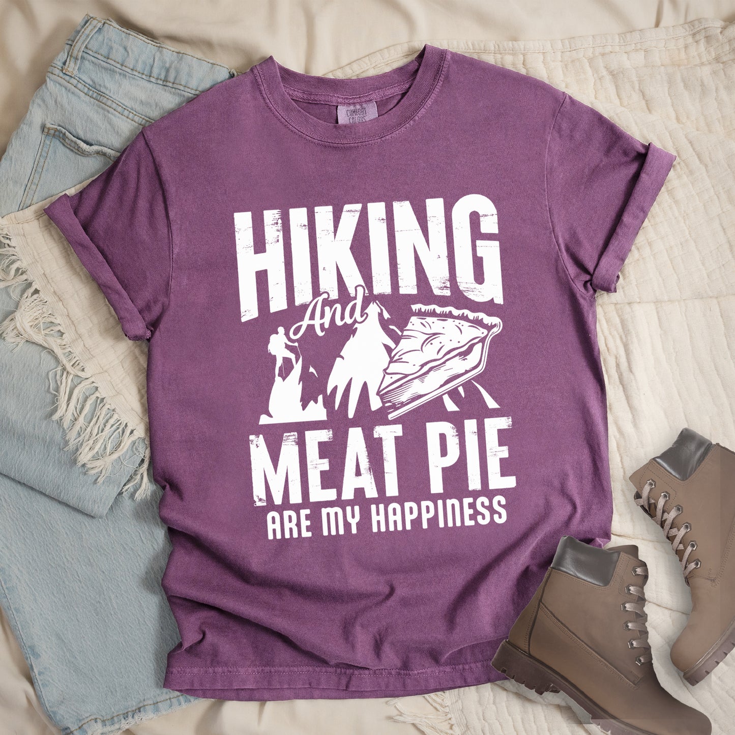 A Berry (Dark Violet) t-shirt featuring white distressed text reading "HIKING And MEAT PIE ARE MY HAPPINESS." Between the words "HIKING" and "MEAT PIE" is a graphic showing a mountain silhouette with a tiny hiker climbing it, alongside an illustration of a slice of meat pie. The design uses only white graphics and text on the Berry (Dark Violet) shirt background in a rugged, vintage style. Features a classic crew neck and casual rolled sleeves, styled alongside light wash denim and brown hiking boots.