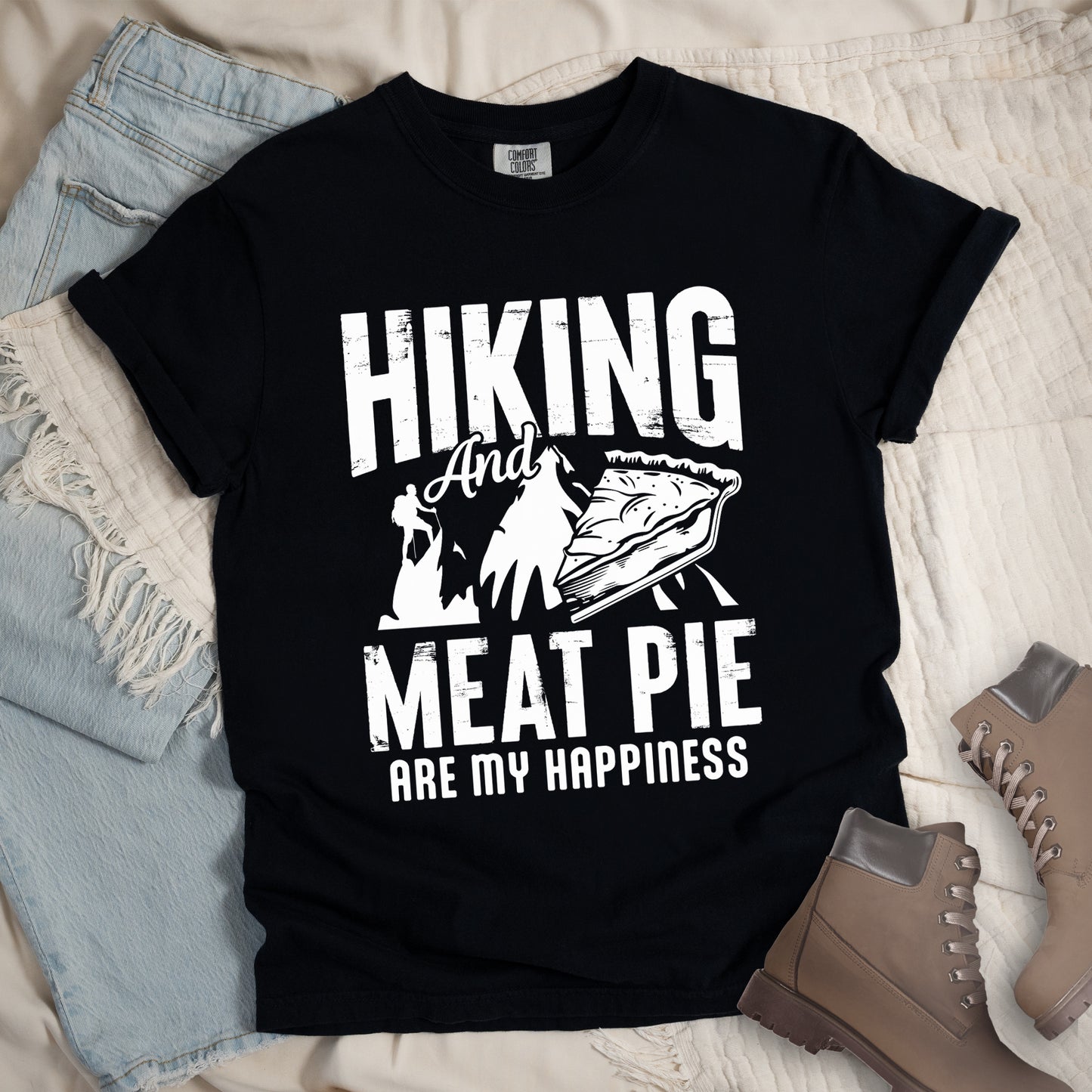 A black t-shirt featuring white distressed text reading "HIKING And MEAT PIE ARE MY HAPPINESS." Between the words "HIKING" and "MEAT PIE" is a graphic showing a mountain silhouette with a tiny hiker climbing it, alongside an illustration of a slice of meat pie. The design uses only white graphics and text on the black shirt background in a rugged, vintage style. The shirt features a classic crew neck and casual rolled sleeves, styled alongside light wash denim and brown hiking boots.