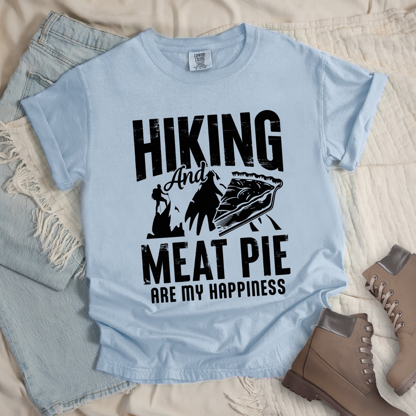 A Chambray (Light Blue) t-shirt featuring black distressed text reading "HIKING And MEAT PIE ARE MY HAPPINESS." Between the words "HIKING" and "MEAT PIE" is a graphic showing a mountain silhouette with a tiny hiker climbing it, alongside an illustration of a slice of meat pie. The design uses only black graphics and text on the Chambray (Light Blue) shirt background in a rugged, vintage style. Features a classic crew neck and casual rolled sleeves, styled alongside light wash denim and brown hiking boots.