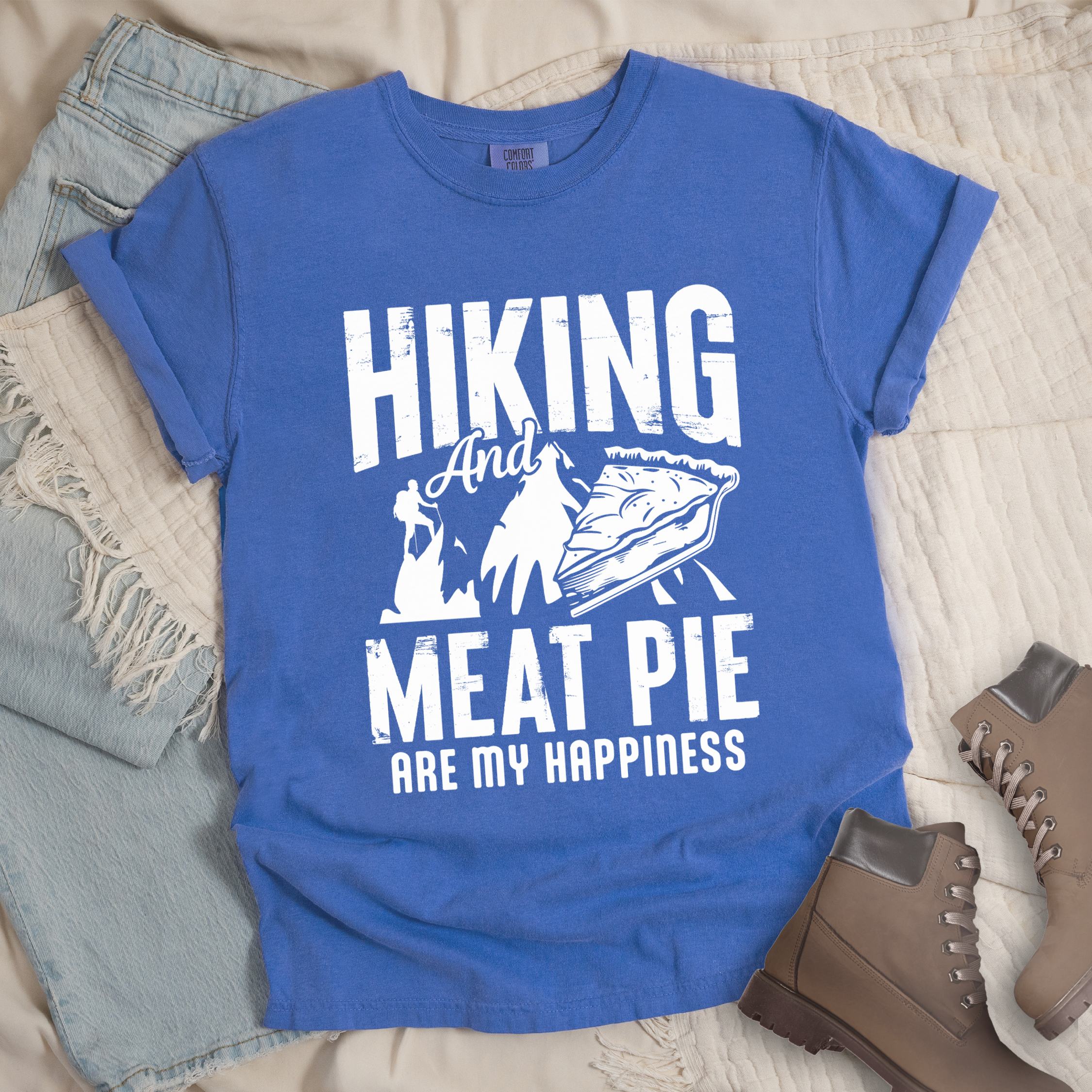 Blue shirt showing a hiking design with 'Hiking and meat pie are my happiness' phrase in front of a mountain and hiker silhouette. In front a slice of pie is shown.