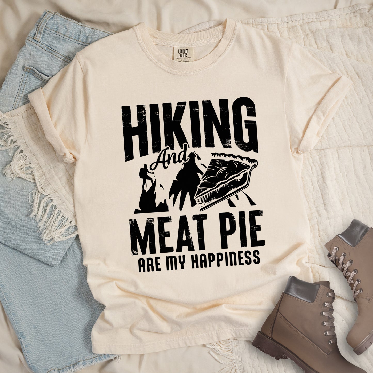 An Ivory (Cream White) t-shirt featuring black distressed text reading "HIKING And MEAT PIE ARE MY HAPPINESS." Between the words "HIKING" and "MEAT PIE" is a graphic showing a mountain silhouette with a tiny hiker climbing it, alongside an illustration of a slice of meat pie. The design uses only black graphics and text on the Ivory (Cream White) shirt background in a rugged, vintage style. Features a classic crew neck and casual rolled sleeves, styled alongside light wash denim and brown hiking boots.