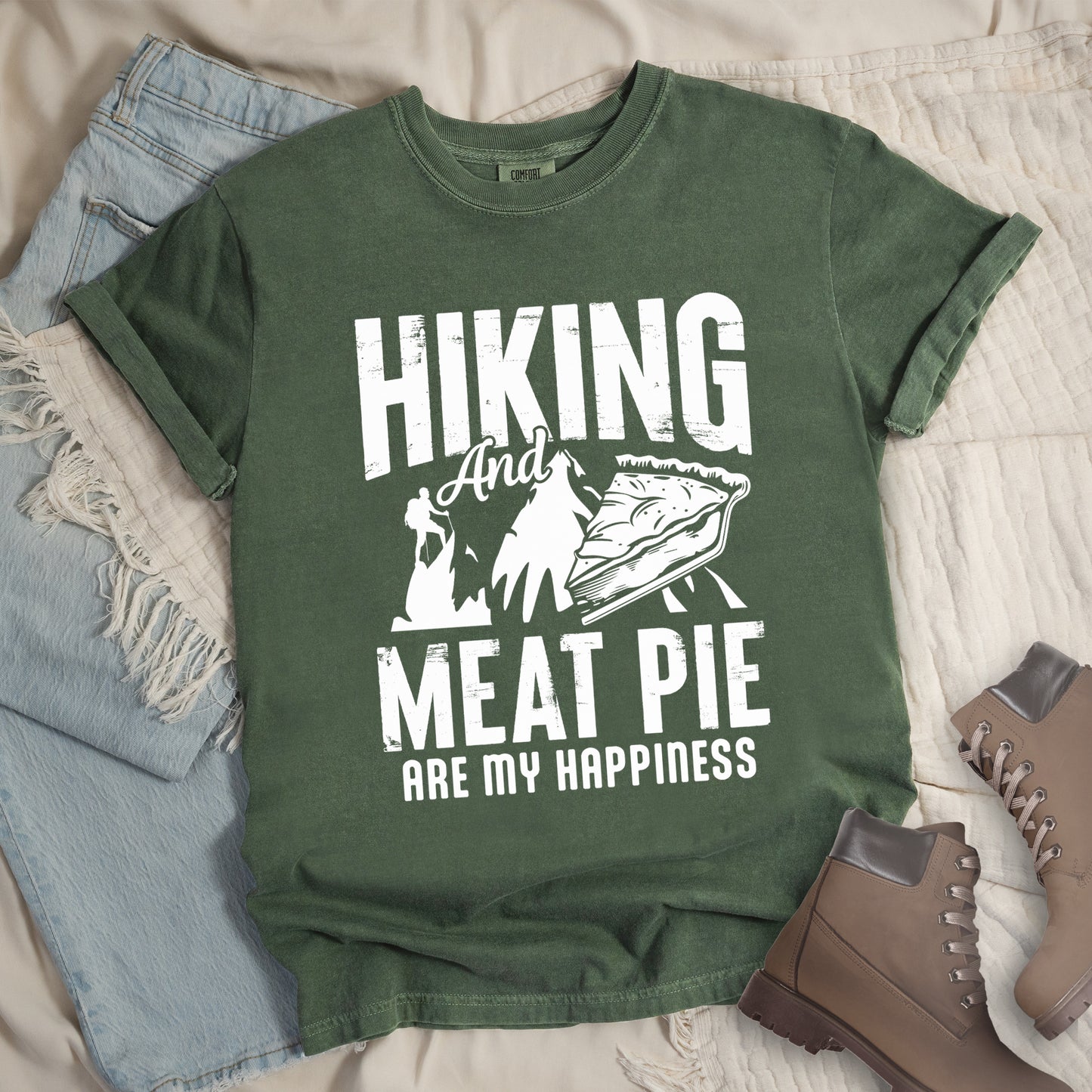 A Moss (Dark Green) t-shirt featuring white distressed text reading "HIKING And MEAT PIE ARE MY HAPPINESS." Between the words "HIKING" and "MEAT PIE" is a graphic showing a mountain silhouette with a tiny hiker climbing it, alongside an illustration of a slice of meat pie. The design uses only white graphics and text on the Moss (Dark Green) shirt background in a rugged, vintage style. Features a classic crew neck and casual rolled sleeves, styled alongside light wash denim and brown hiking boots.