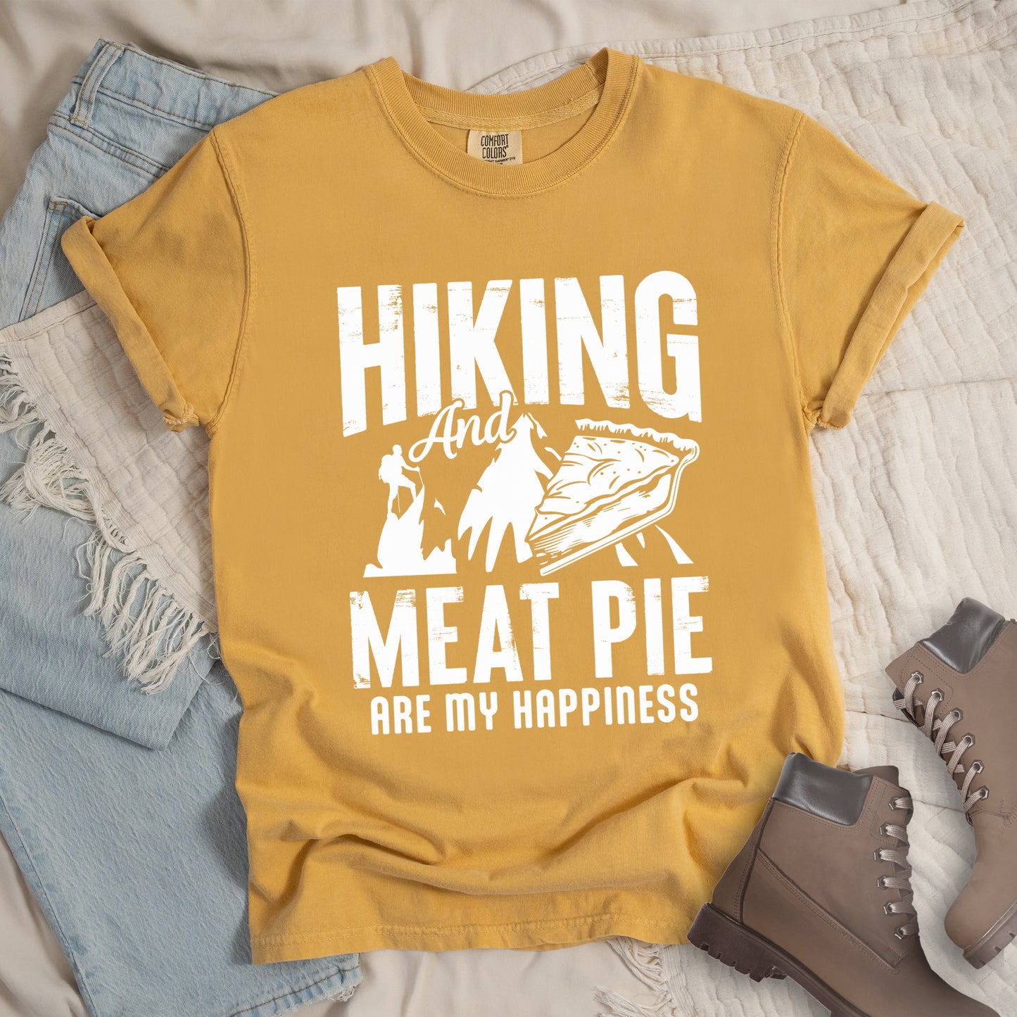A Mustard (Yellow) t-shirt featuring white distressed text reading "HIKING And MEAT PIE ARE MY HAPPINESS." Between the words "HIKING" and "MEAT PIE" is a graphic showing a mountain silhouette with a tiny hiker climbing it, alongside an illustration of a slice of meat pie. The design uses only white graphics and text on the Mustard (Yellow) shirt background in a rugged, vintage style. Features a classic crew neck and casual rolled sleeves, styled alongside light wash denim and brown hiking boots.