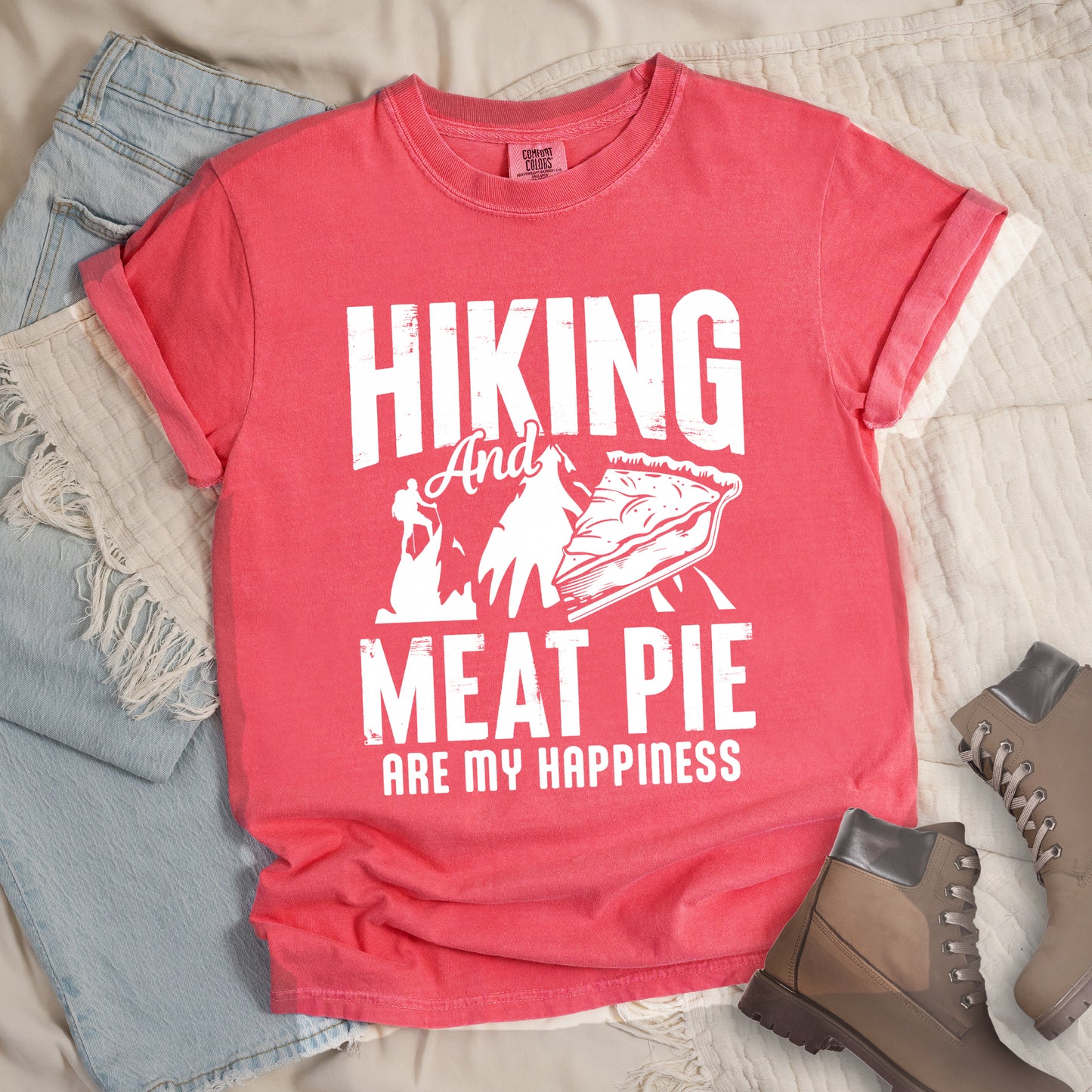 A Watermelon (Light Coral) t-shirt featuring white distressed text reading "HIKING And MEAT PIE ARE MY HAPPINESS." Between the words "HIKING" and "MEAT PIE" is a graphic showing a mountain silhouette with a tiny hiker climbing it, alongside an illustration of a slice of meat pie. The design uses only white graphics and text on the Watermelon shirt background in a rugged, vintage style. Features a classic crew neck and casual rolled sleeves, styled alongside light wash denim and brown hiking boots.