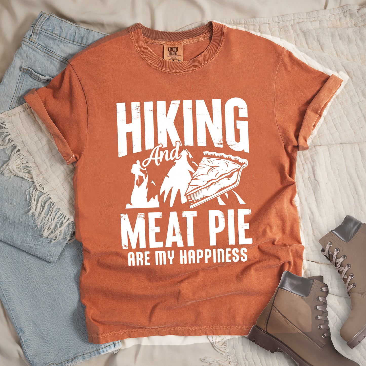 A Yam (Brown) t-shirt featuring white distressed text reading "HIKING And MEAT PIE ARE MY HAPPINESS." Between the words "HIKING" and "MEAT PIE" is a graphic showing a mountain silhouette with a tiny hiker climbing it, alongside an illustration of a slice of meat pie. The design uses only white graphics and text on the Yam (Brown) shirt background in a rugged, vintage style. Features a classic crew neck and casual rolled sleeves, styled alongside light wash denim and brown hiking boots.