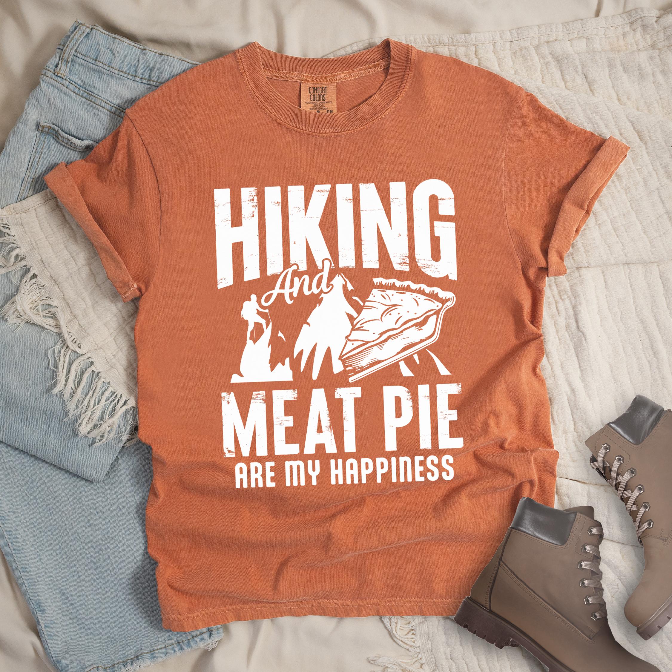 Orange shirt showing a hiking design with 'Hiking and meat pie are my happiness' phrase in front of a mountain and hiker silhouette. In front a slice of pie is shown.