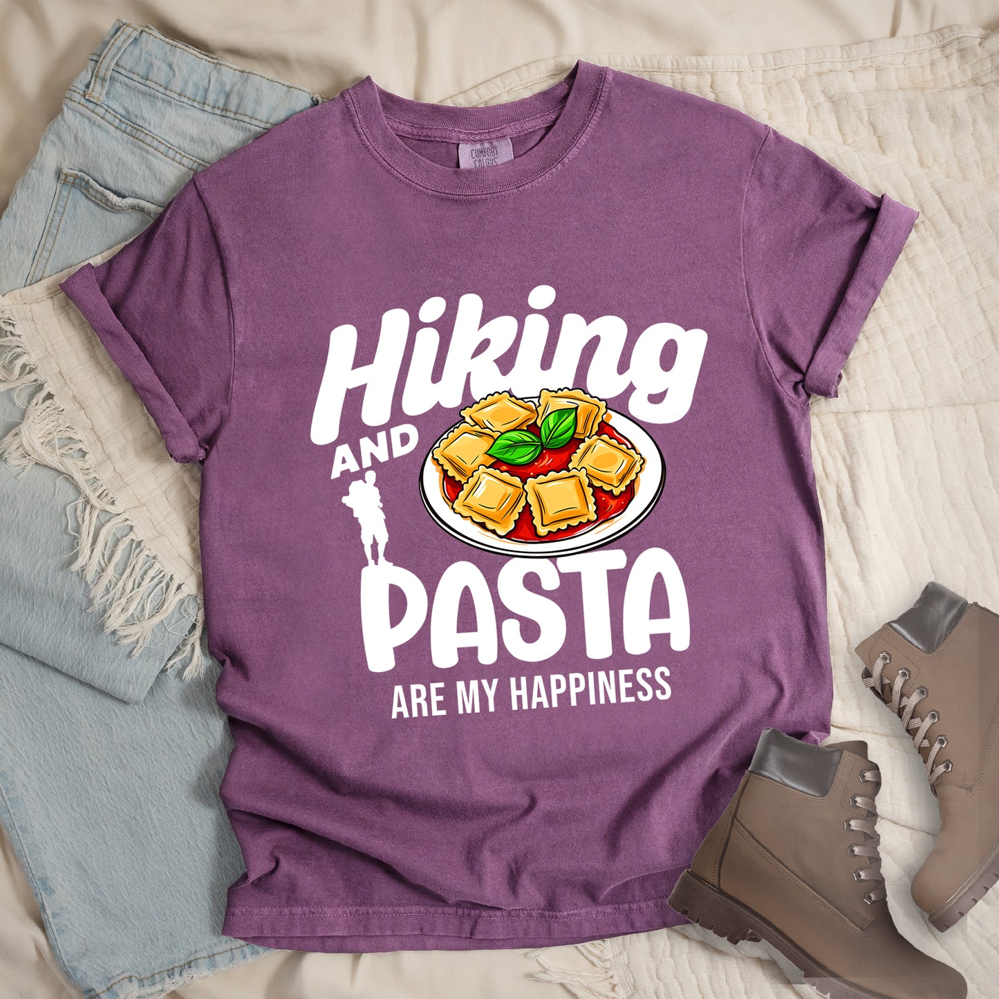 Berry (Dark Violet) t-shirt with text "Hiking AND PASTA ARE MY HAPPINESS". "Hiking" appears in stylized white text at the top, "AND" in smaller white text, "PASTA" in large white letters, and "ARE MY HAPPINESS" in small white text at the bottom. Features a white silhouette of a hiker with backpack on the left and an illustration of ravioli pasta with tomato sauce and basil leaves on a white plate. Features a classic crew neck and casual rolled sleeves, alongside light wash denim and brown hiking boots.