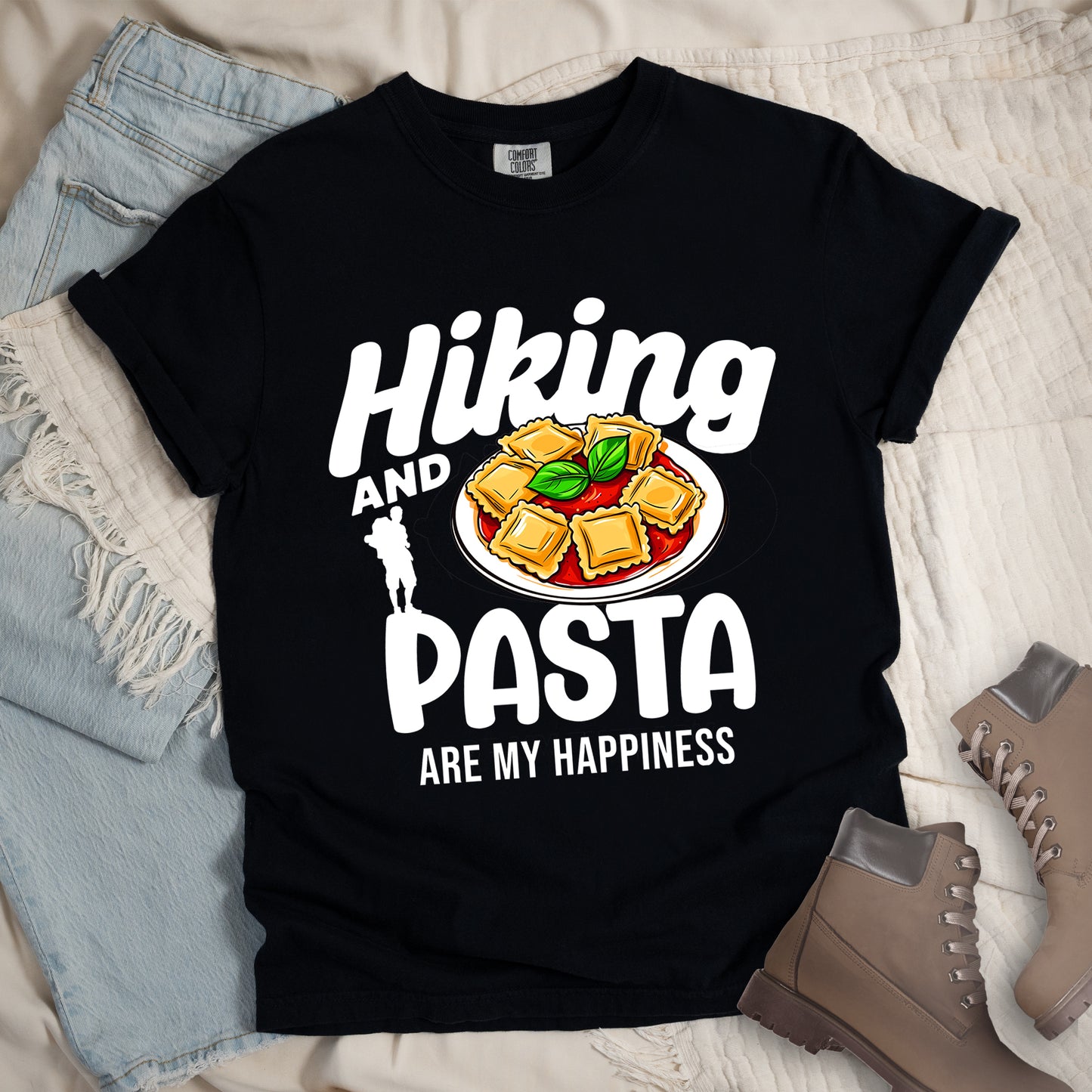 Black t-shirt with white text reading "Hiking AND PASTA ARE MY HAPPINESS". "Hiking" appears in stylized white text at the top, "AND" in smaller white text, "PASTA" in large white letters, and "ARE MY HAPPINESS" in small white text at the bottom. Features a white silhouette of a hiker with backpack on the left and an illustration of ravioli pasta with tomato sauce and basil leaves on a white plate. Features a classic crew neck and casual rolled sleeves, alongside light wash denim and brown hiking boots.