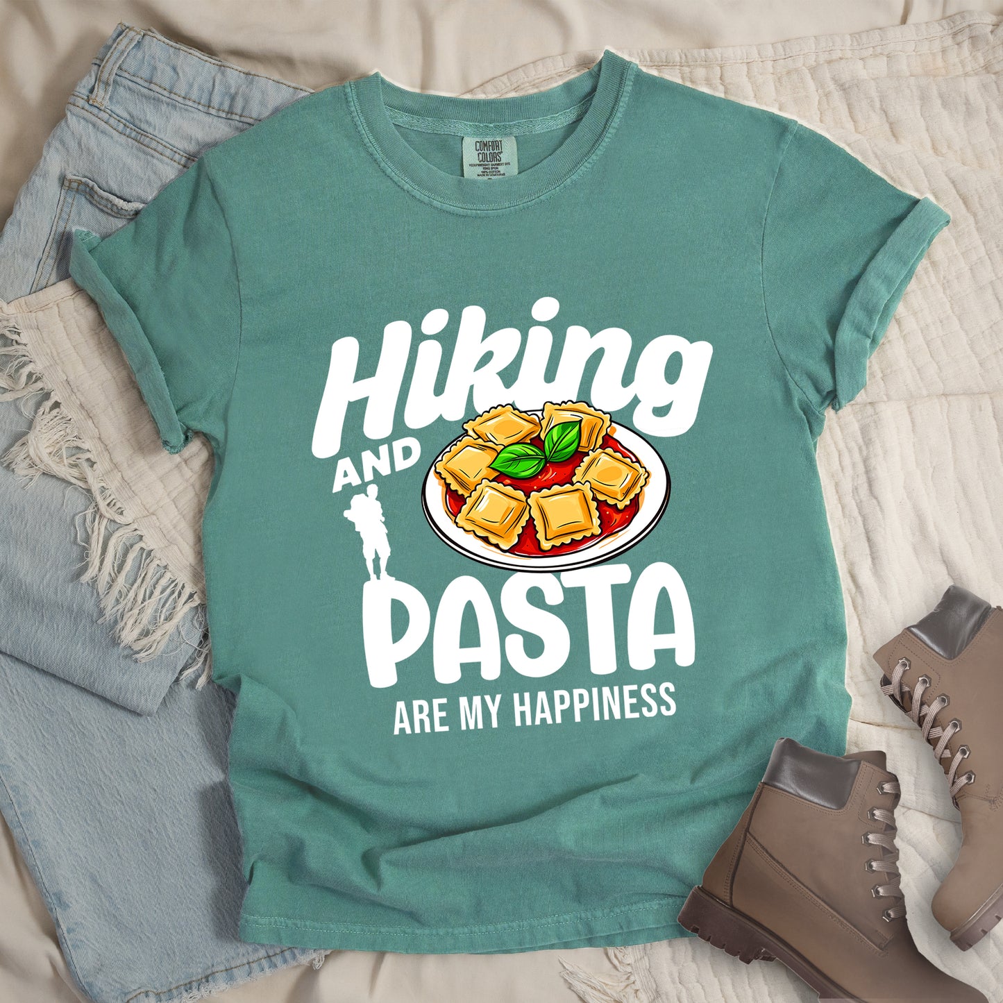 Light Green t-shirt with white text reading "Hiking AND PASTA ARE MY HAPPINESS". "Hiking" appears in stylized white text at the top, "AND" in smaller white text, "PASTA" in large white letters, and "ARE MY HAPPINESS" in small white text at the bottom. Features a white silhouette of a hiker with backpack on the left and an illustration of ravioli pasta with tomato sauce and basil leaves on a white plate. Features a classic crew neck and casual rolled sleeves, alongside light wash denim and brown hiking boots