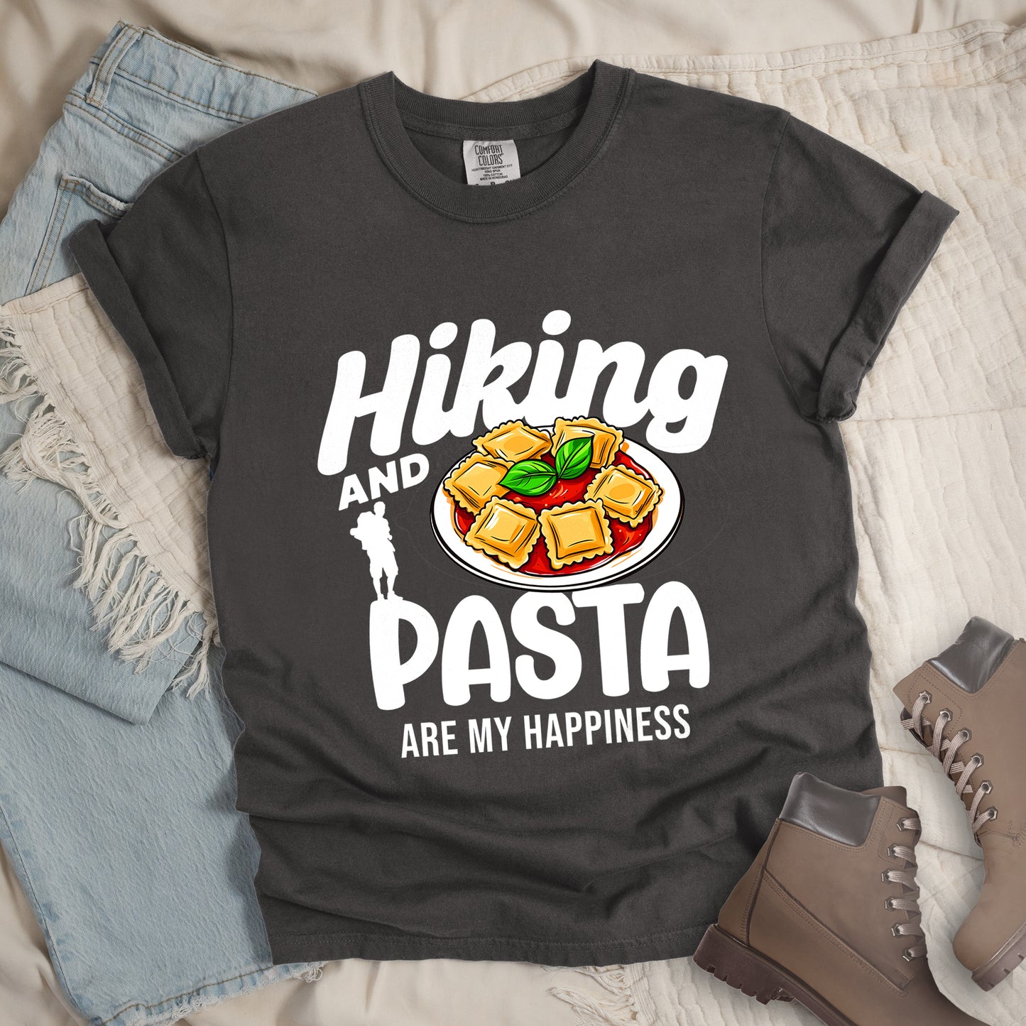 Pepper (Dark Gray) t-shirt with text "Hiking AND PASTA ARE MY HAPPINESS". "Hiking" appears in stylized white text at the top, "AND" in smaller white text, "PASTA" in large white letters, and "ARE MY HAPPINESS" in small white text at the bottom. Features a white silhouette of a hiker with backpack on the left and an illustration of ravioli pasta with tomato sauce and basil leaves on a white plate. Features a classic crew neck and casual rolled sleeves, alongside light wash denim and brown hiking boots.
