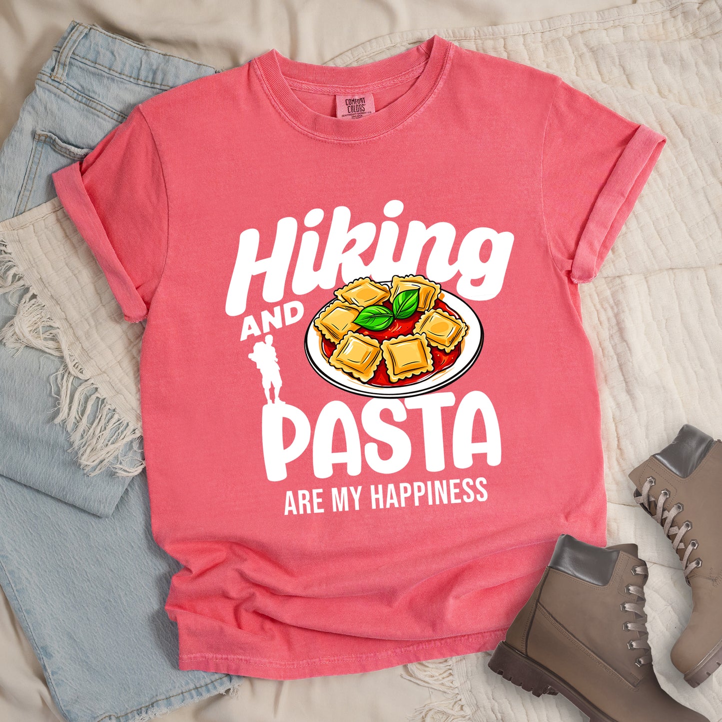 Watermelon (Light Coral) t-shirt with text "Hiking AND PASTA ARE MY HAPPINESS". "Hiking" appears in stylized white text at the top, "AND" in smaller white text, "PASTA" in large white letters, and "ARE MY HAPPINESS" in small white text at the bottom. Features a white silhouette of a hiker with backpack on the left and an illustration of ravioli pasta with tomato sauce and basil leaves on a white plate. Features a classic crew neck and casual rolled sleeves, alongside light wash denim and brown hiking boots