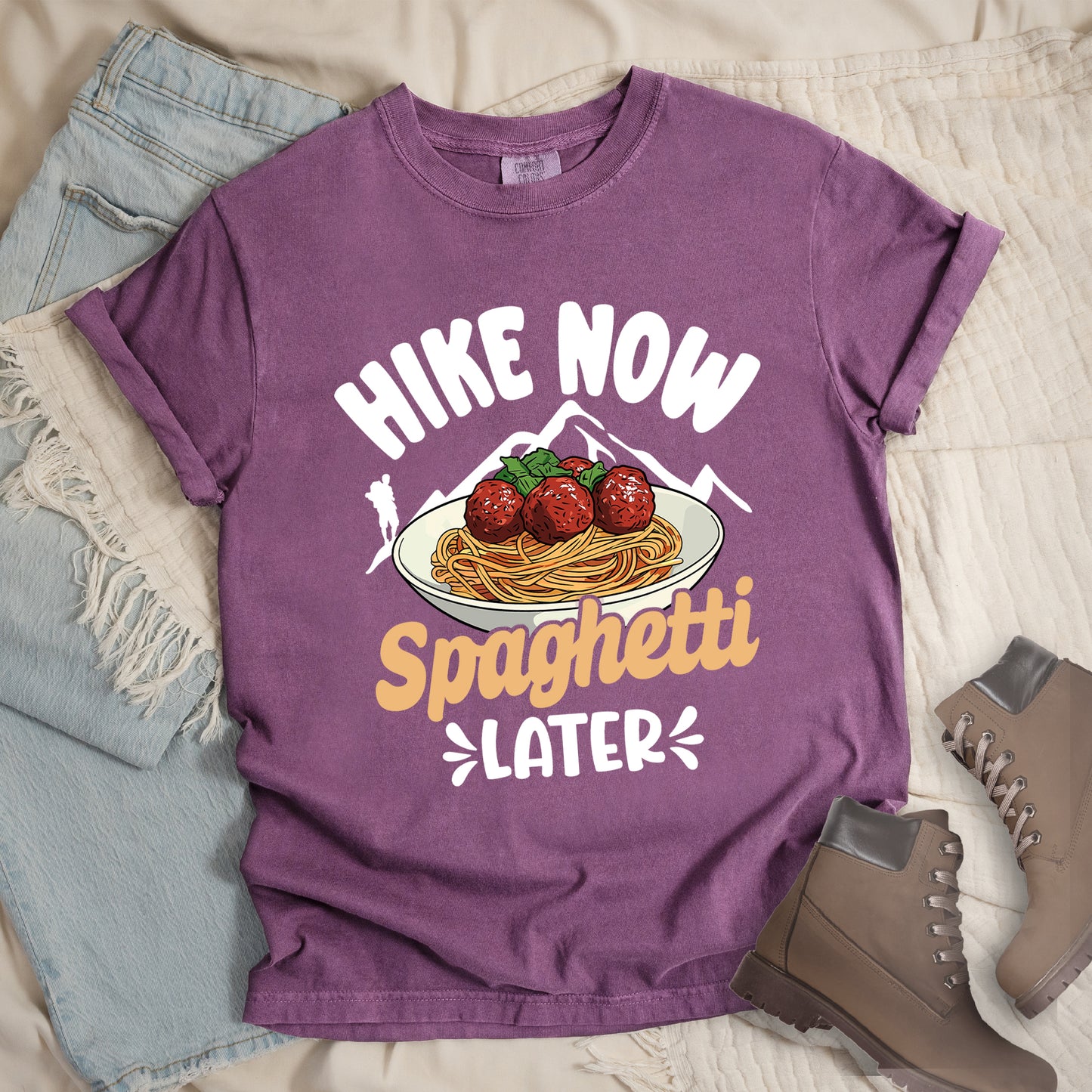 Berry (Dark Violet) t-shirt with white and orange text reading "HIKE NOW Spaghetti LATER". "HIKE NOW" appears in curved white text at the top, "Spaghetti" in orange script in the middle, and "LATER" in white at the bottom with small decorative accents. Features white mountain silhouettes with a white hiker silhouette on the left, and a detailed illustration of spaghetti and meatballs with basil on a white plate.
