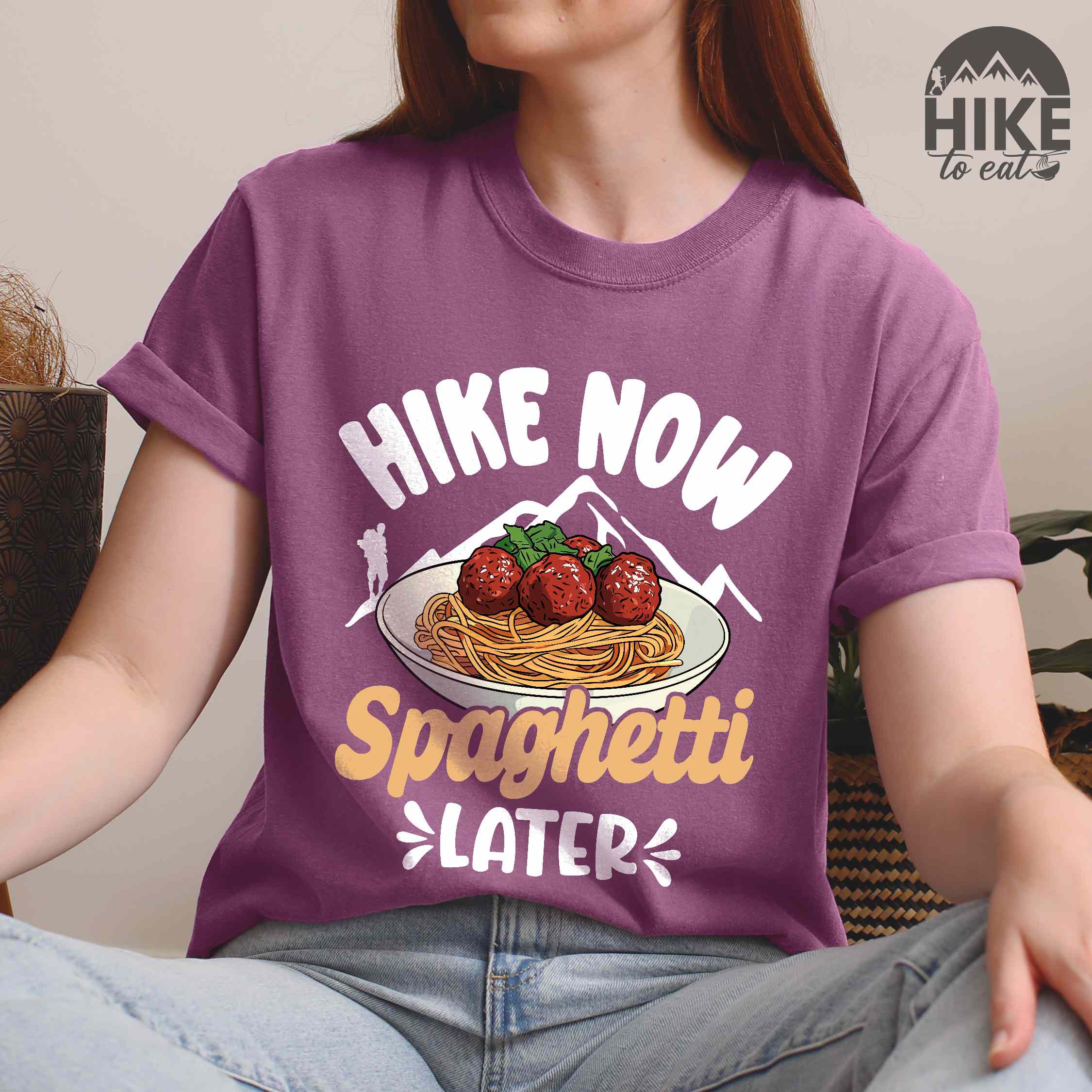 Woman wearing berry "Hike now spaghetti later" colored vintage tee with bold typography, mountains, birds, illustrated plate of spaghetti meatballs, and backpackers for pasta lovers.