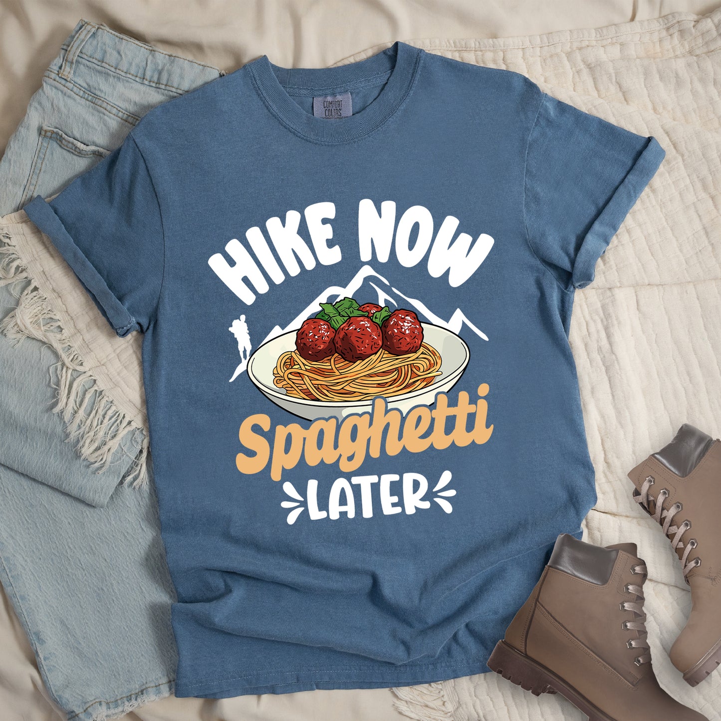 Blue Jean (Denim Blue) t-shirt with white and orange text reading "HIKE NOW Spaghetti LATER". "HIKE NOW" appears in curved white text at the top, "Spaghetti" in orange script in the middle, and "LATER" in white at the bottom with small decorative accents. Features white mountain silhouettes with a white hiker silhouette on the left, and a detailed illustration of spaghetti and meatballs with basil on a white plate.