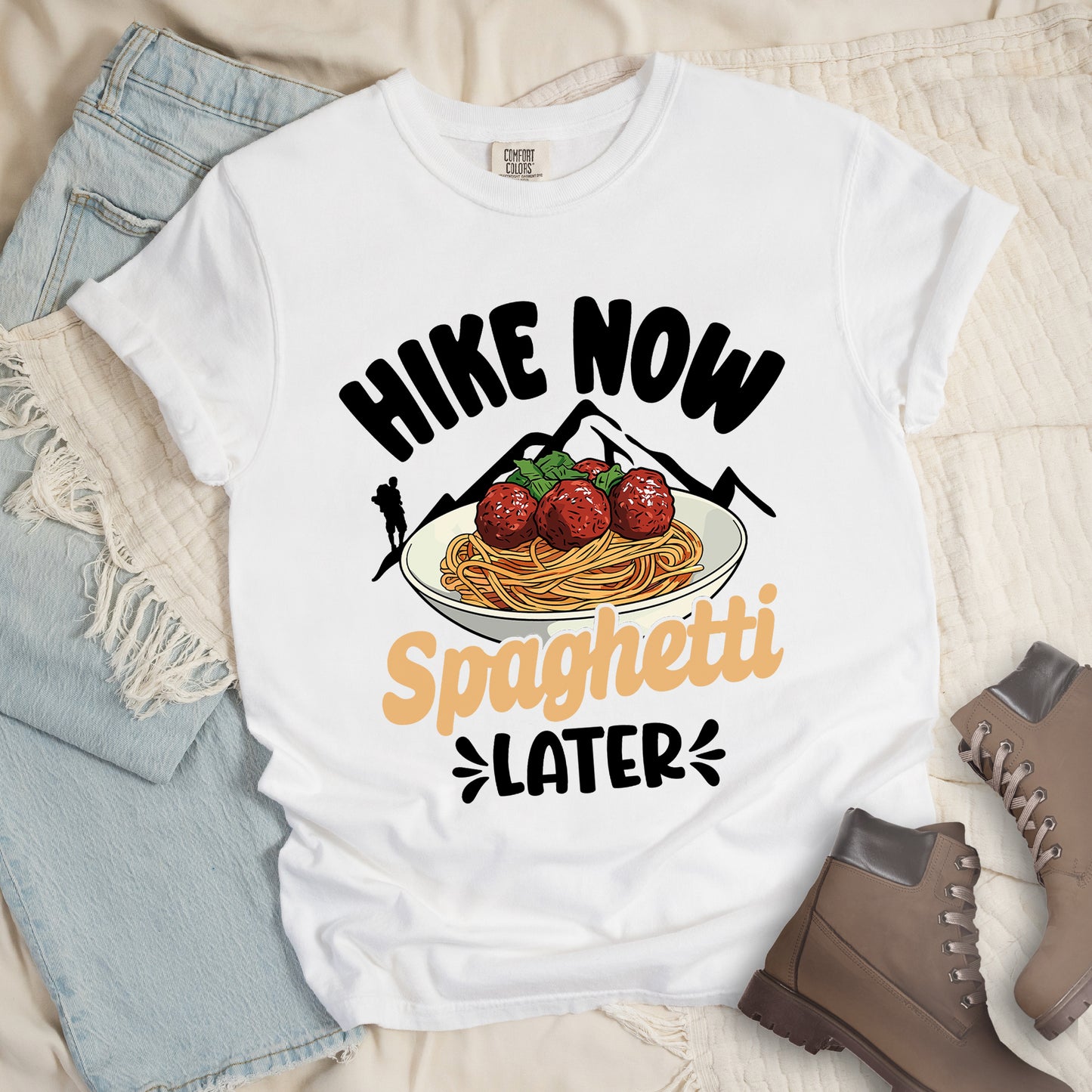 White t-shirt with black and orange text reading "HIKE NOW Spaghetti LATER". "HIKE NOW" appears in curved black text at the top, "Spaghetti" in orange script in the middle, and "LATER" in black at the bottom with small decorative accents. Features black mountain silhouettes with a black hiker silhouette on the left, and a detailed illustration of spaghetti and meatballs with basil on a white plate.