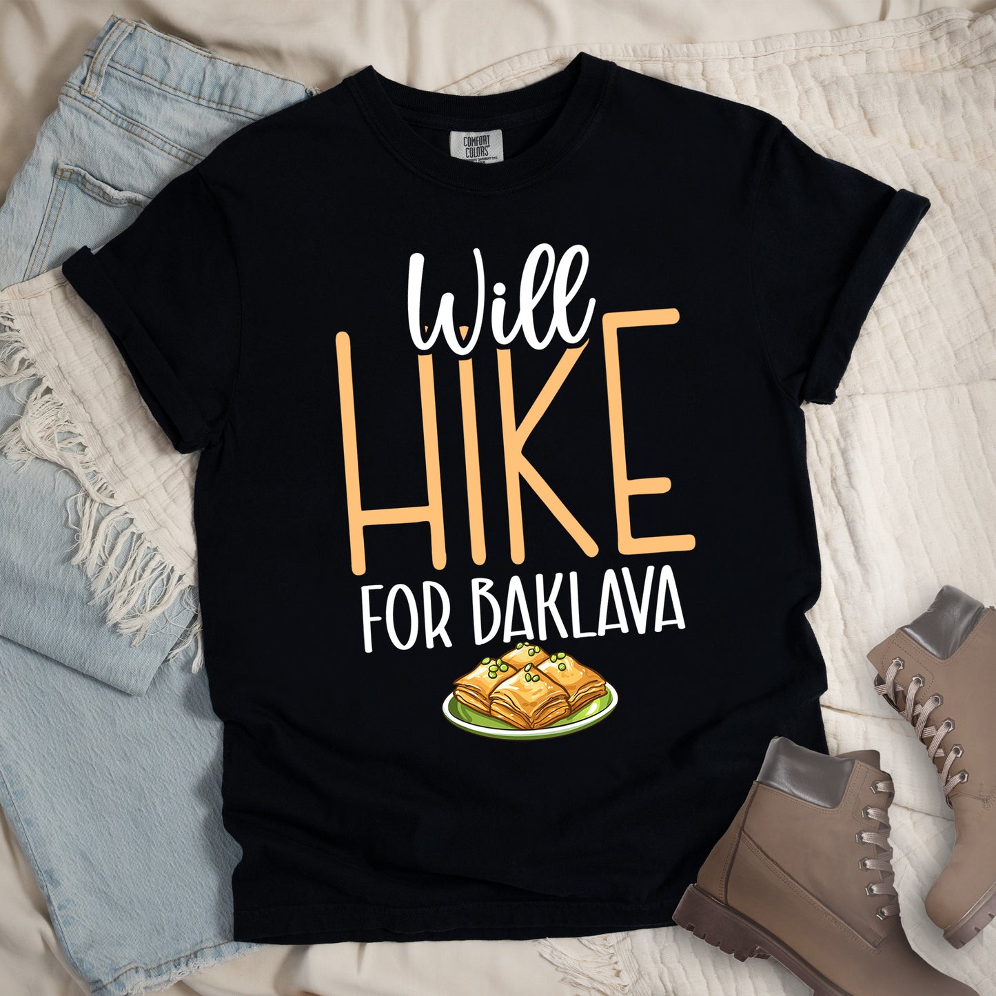 Black t-shirt with white and peach text reading "Will HIKE FOR BAKLAVA". "Will" appears in white script at the top, "HIKE" in large peach letters in the middle, and "FOR BAKLAVA" in white at the bottom. Features an illustration of baklava pieces with honey and pistachios on a green plate below the text. Features a classic crew neck and casual rolled sleeves, styled alongside light wash denim and brown hiking boots.