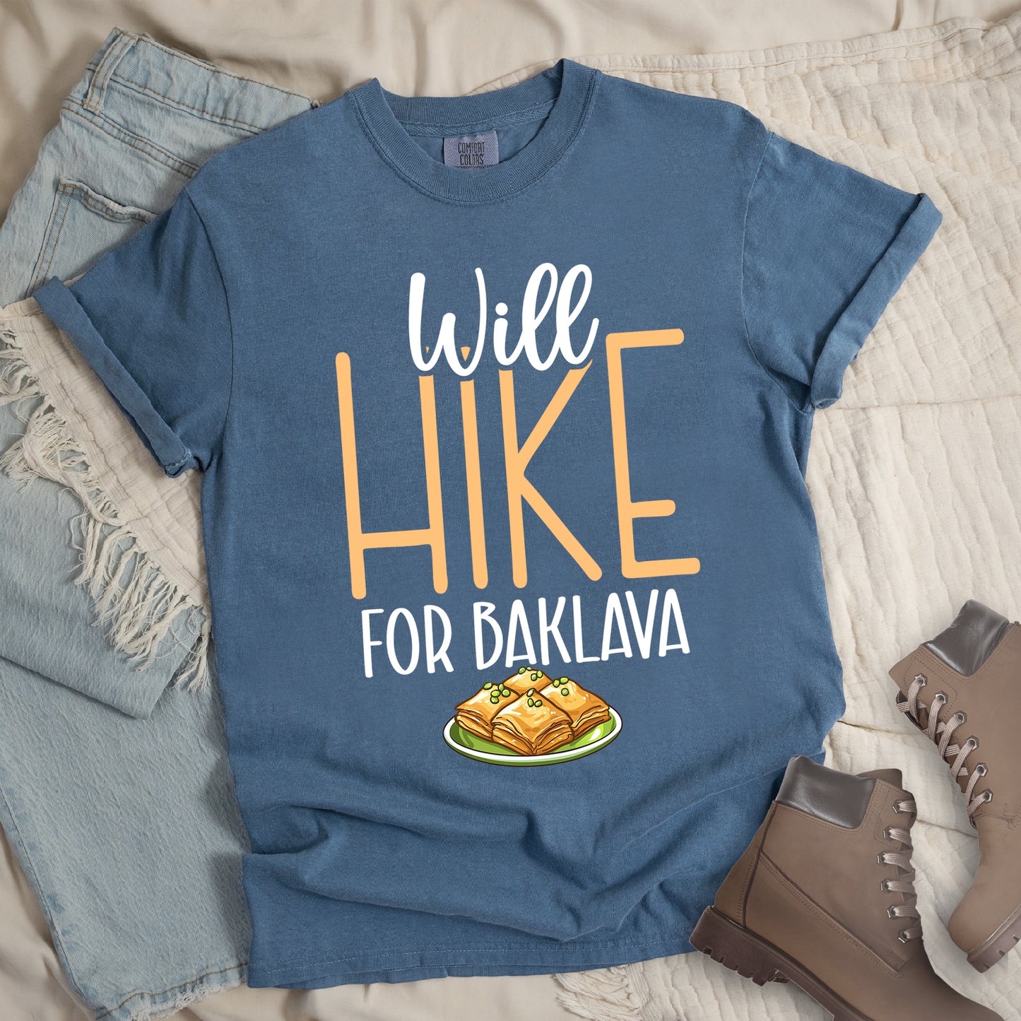Blue Jean (Denim Blue) t-shirt with white and peach text reading "Will HIKE FOR BAKLAVA". "Will" appears in white script at the top, "HIKE" in large peach letters in the middle, and "FOR BAKLAVA" in white at the bottom. Features an illustration of baklava pieces with honey and pistachios on a green plate below the text. Features a classic crew neck and casual rolled sleeves, styled alongside light wash denim and brown hiking boots.