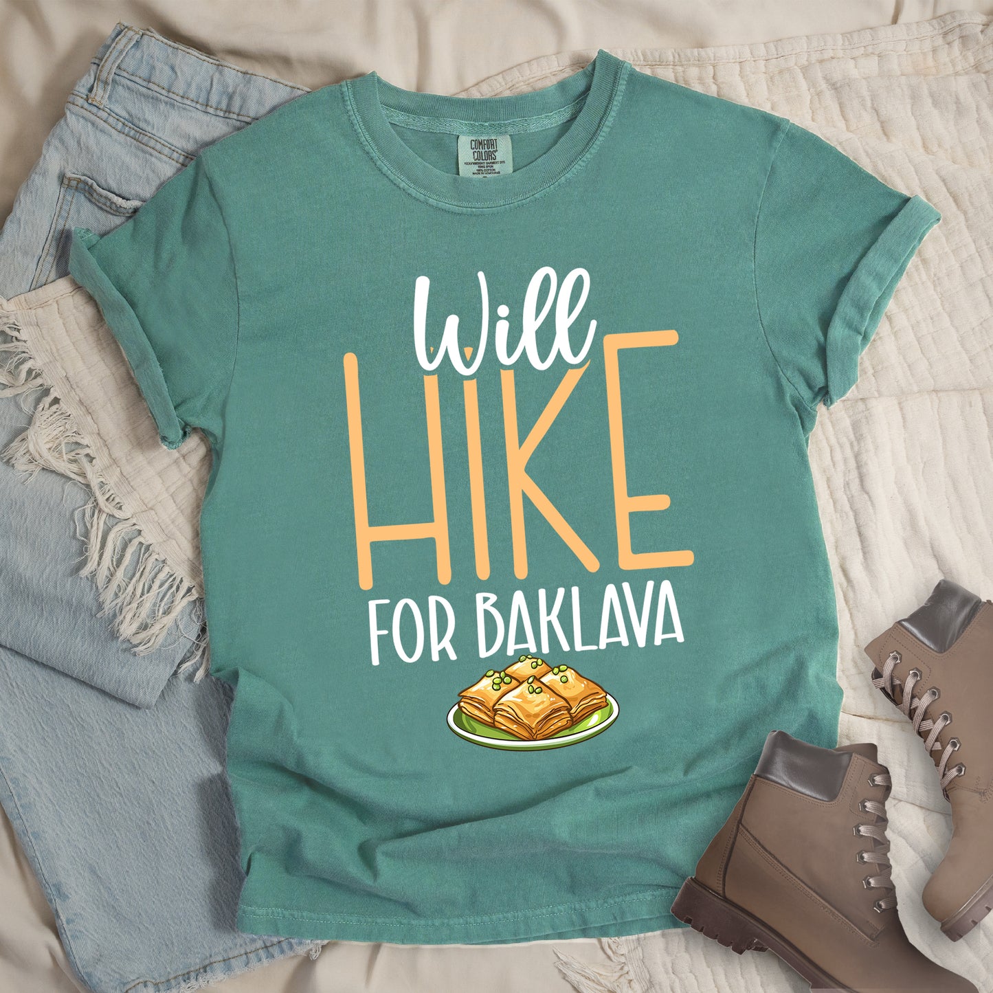 Light Green t-shirt with white and peach text reading "Will HIKE FOR BAKLAVA". "Will" appears in white script at the top, "HIKE" in large peach letters in the middle, and "FOR BAKLAVA" in white at the bottom. Features an illustration of baklava pieces with honey and pistachios on a green plate below the text. Features a classic crew neck and casual rolled sleeves, styled alongside light wash denim and brown hiking boots.