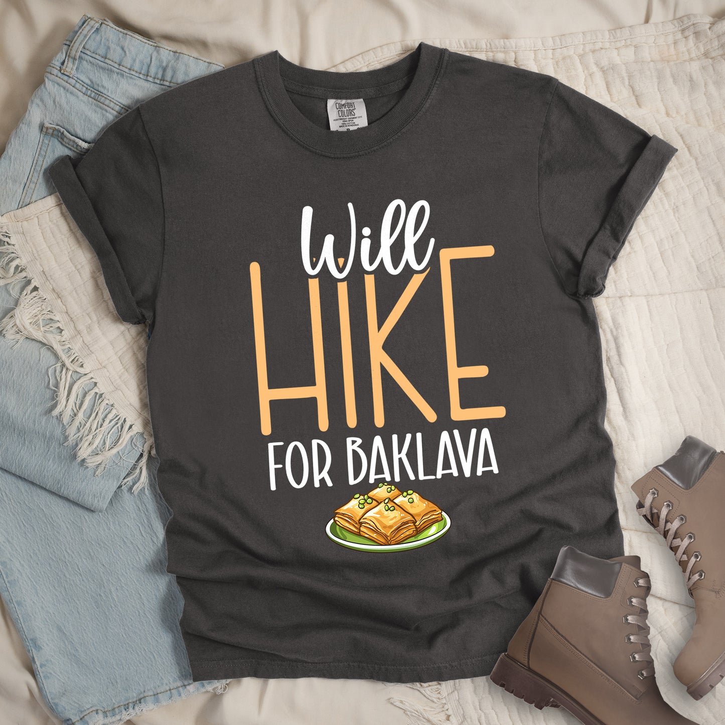Pepper (Dark Gray) t-shirt with white and peach text reading "Will HIKE FOR BAKLAVA". "Will" appears in white script at the top, "HIKE" in large peach letters in the middle, and "FOR BAKLAVA" in white at the bottom. Features an illustration of baklava pieces with honey and pistachios on a green plate below the text. Features a classic crew neck and casual rolled sleeves, styled alongside light wash denim and brown hiking boots.