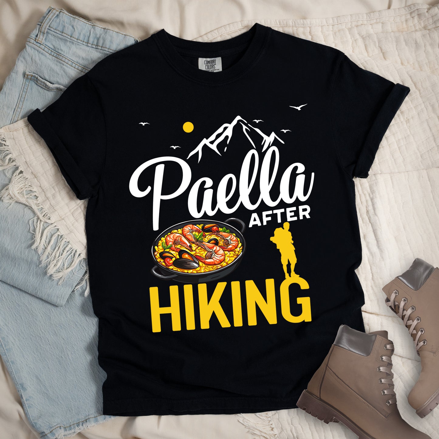 Black t-shirt with white and yellow text reading "Paella AFTER HIKING". "Paella" appears in white script at the top, "AFTER" in white text in the middle, and "HIKING" in large yellow letters at the bottom. Features white mountain silhouettes, birds, and a yellow sun. A yellow hiker silhouette appears between "AFTER" and on top of the letter "G" in "HIKING" on the right, with a detailed illustration of a paella dish with seafood in a black pan to the left of the hiker.