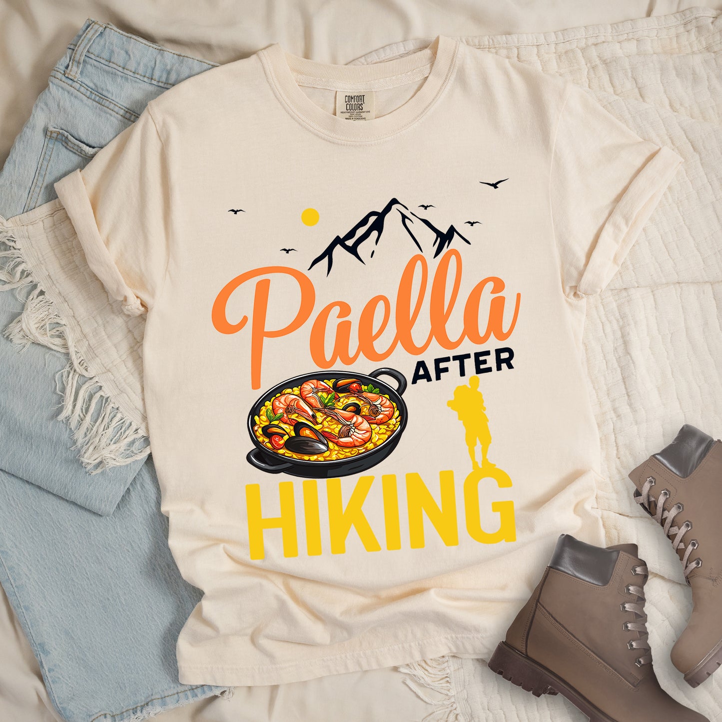 Ivory (Cream White) t-shirt with black, orange and yellow text reading "Paella AFTER HIKING". "Paella" appears in orange script at the top, "AFTER" in black text in the middle, and "HIKING" in large yellow letters at the bottom. Features black mountain silhouettes, birds, and a yellow sun. A yellow hiker silhouette appears between "AFTER" and on top of the letter "G" in "HIKING" on the right, with a detailed illustration of a paella dish with seafood in a black pan to the left of the hiker.