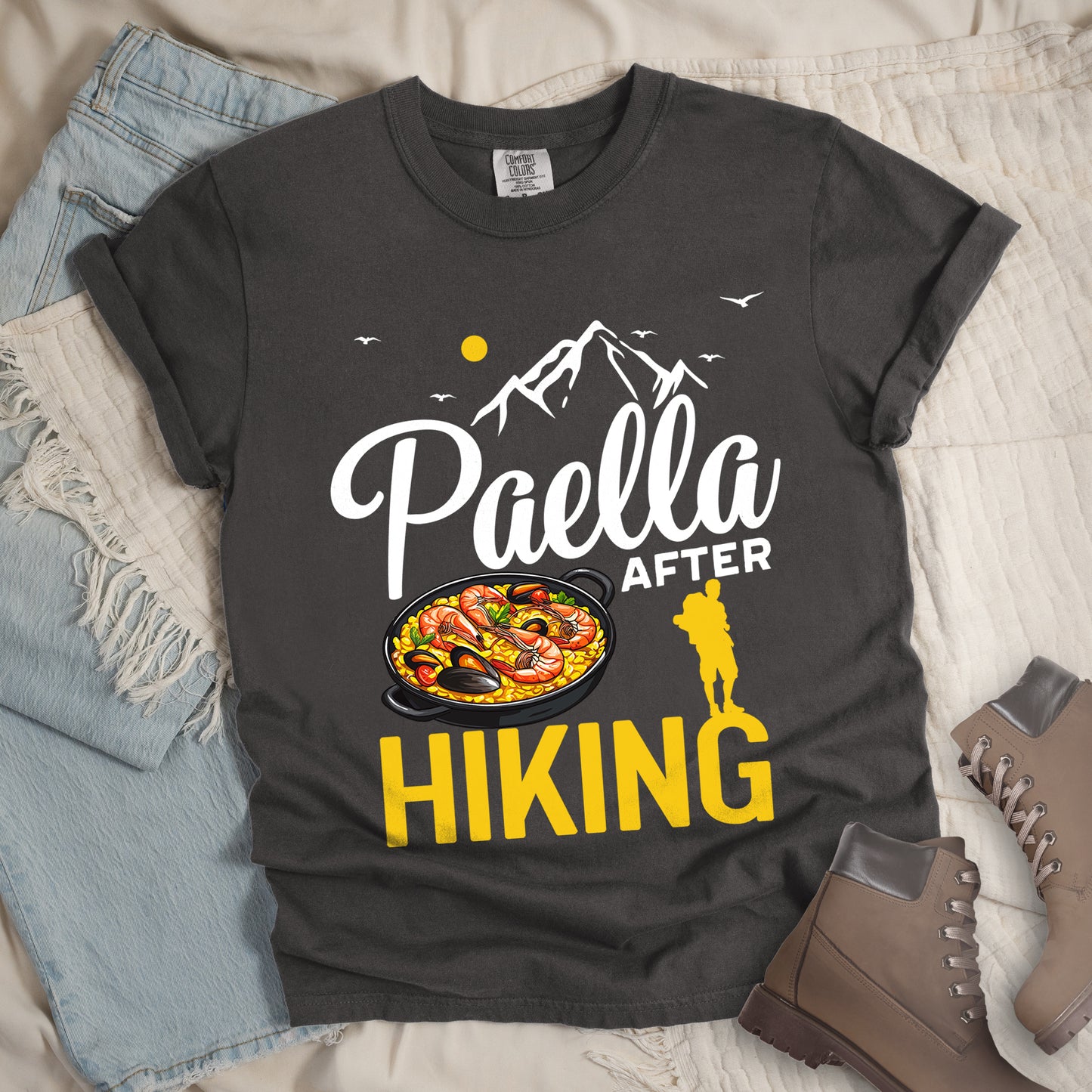 Pepper (Dark Gray) t-shirt with white and yellow text reading "Paella AFTER HIKING". "Paella" appears in white script at the top, "AFTER" in white text in the middle, and "HIKING" in large yellow letters at the bottom. Features white mountain silhouettes, birds, and a yellow sun. A yellow hiker silhouette appears between "AFTER" and on top of the letter "G" in "HIKING" on the right, with a detailed illustration of a paella dish with seafood in a black pan to the left of the hiker.