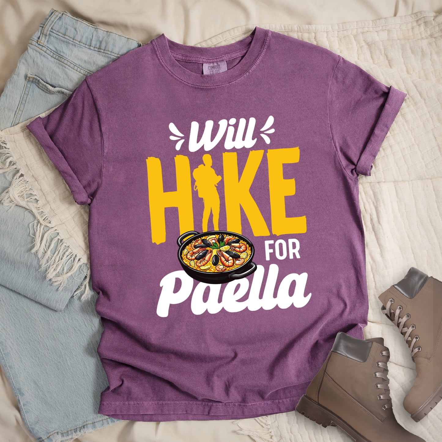 Berry (Dark Violet) t-shirt with white and yellow text reading "Will HIKE FOR Paella". "Will" appears in white script at the top with small decorative accents, "HIKE" in large yellow letters with a yellow hiker silhouette integrated into the letter "I", "FOR" in white, and "Paella" in white script at the bottom. Features an illustration of a paella dish with seafood in a black pan to the left of "FOR" and above "Paella".