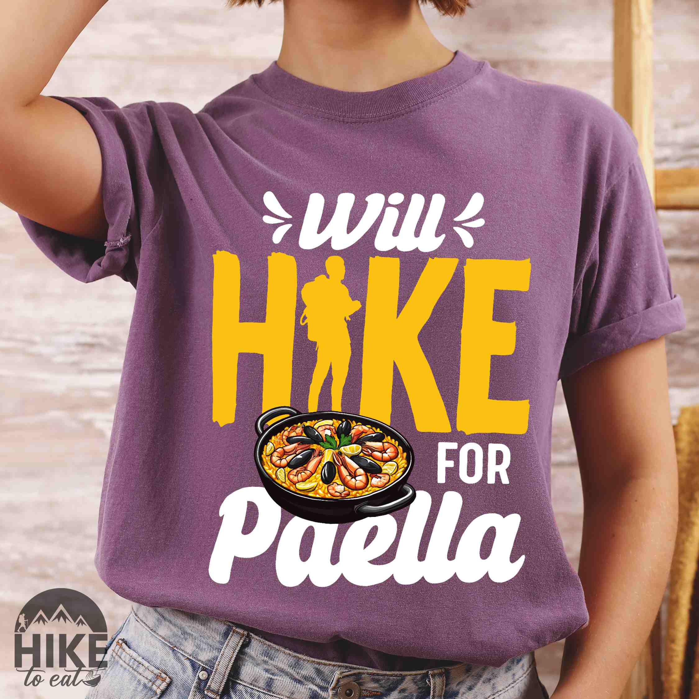 Woman wearing berry "Will Hike for Paella" colored vintage tee with bold typography, illustrated paella platter, and backpackers for rice lovers.