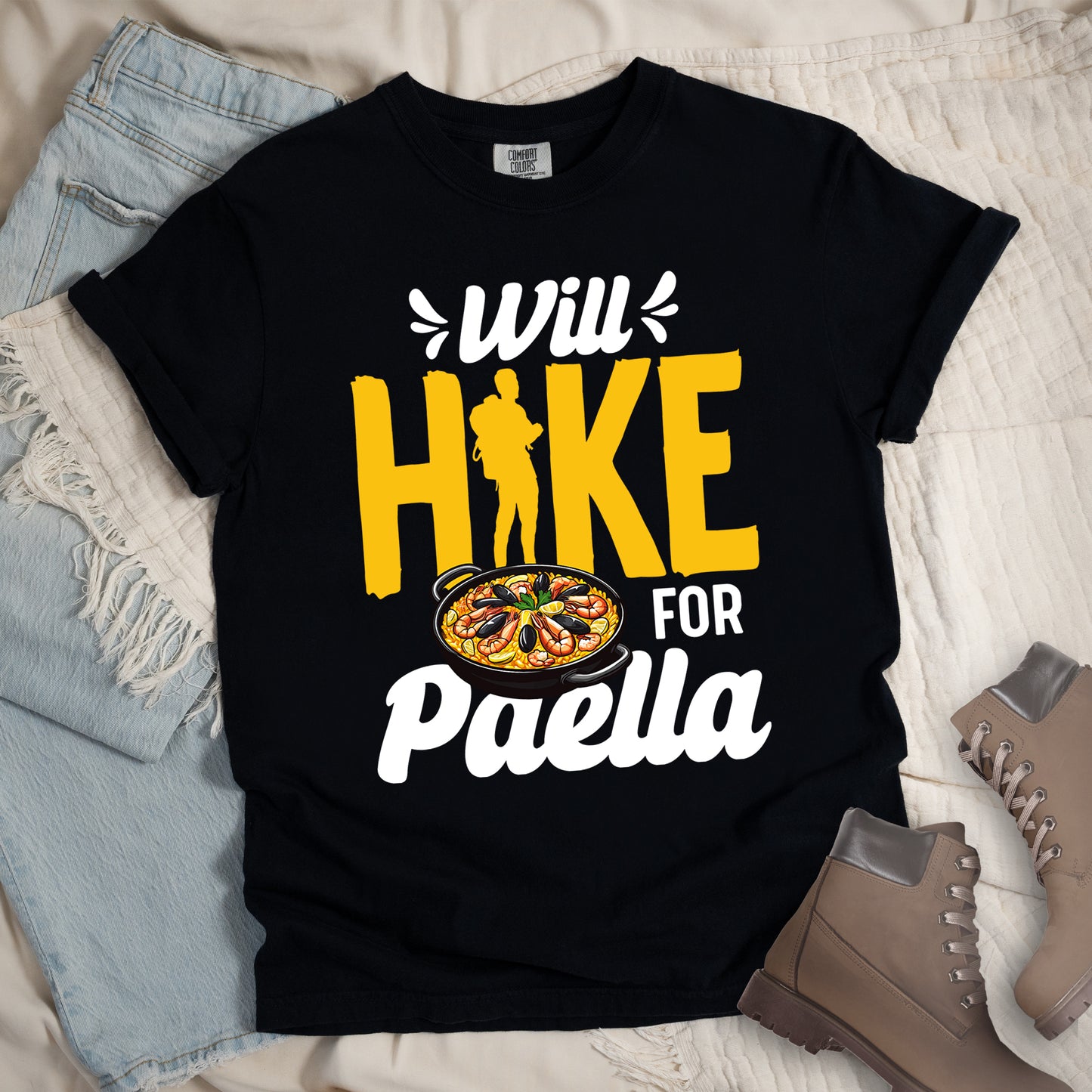 Black t-shirt with white and yellow text reading "Will HIKE FOR Paella". "Will" appears in white script at the top with small decorative accents, "HIKE" in large yellow letters with a yellow hiker silhouette integrated into the letter "I", "FOR" in white, and "Paella" in white script at the bottom. Features an illustration of a paella dish with seafood in a black pan to the left of "FOR" and above "Paella".
