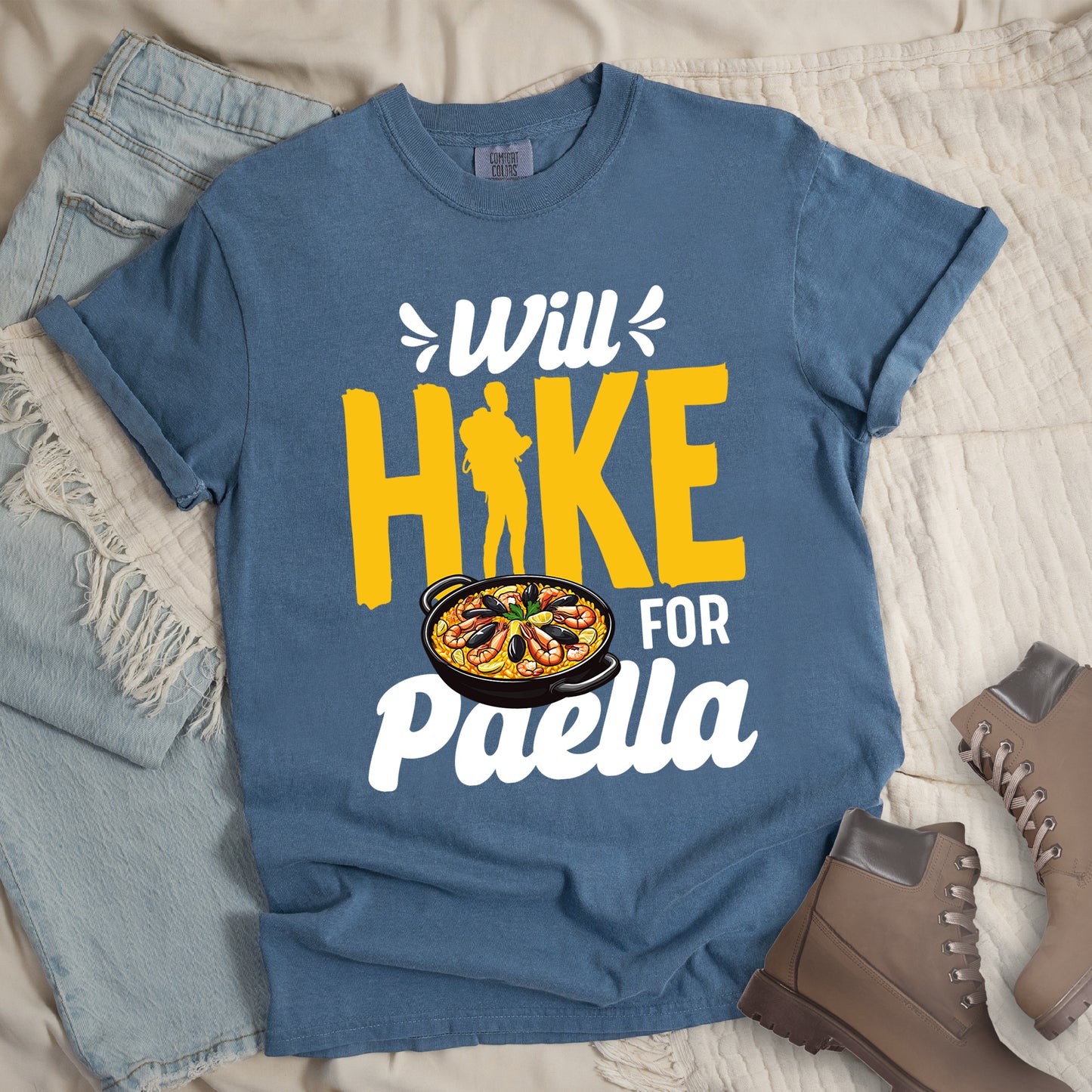 Blue Jean (Denim Blue) t-shirt with white and yellow text reading "Will HIKE FOR Paella". "Will" appears in white script at the top with small decorative accents, "HIKE" in large yellow letters with a yellow hiker silhouette integrated into the letter "I", "FOR" in white, and "Paella" in white script at the bottom. Features an illustration of a paella dish with seafood in a black pan to the left of "FOR" and above "Paella".