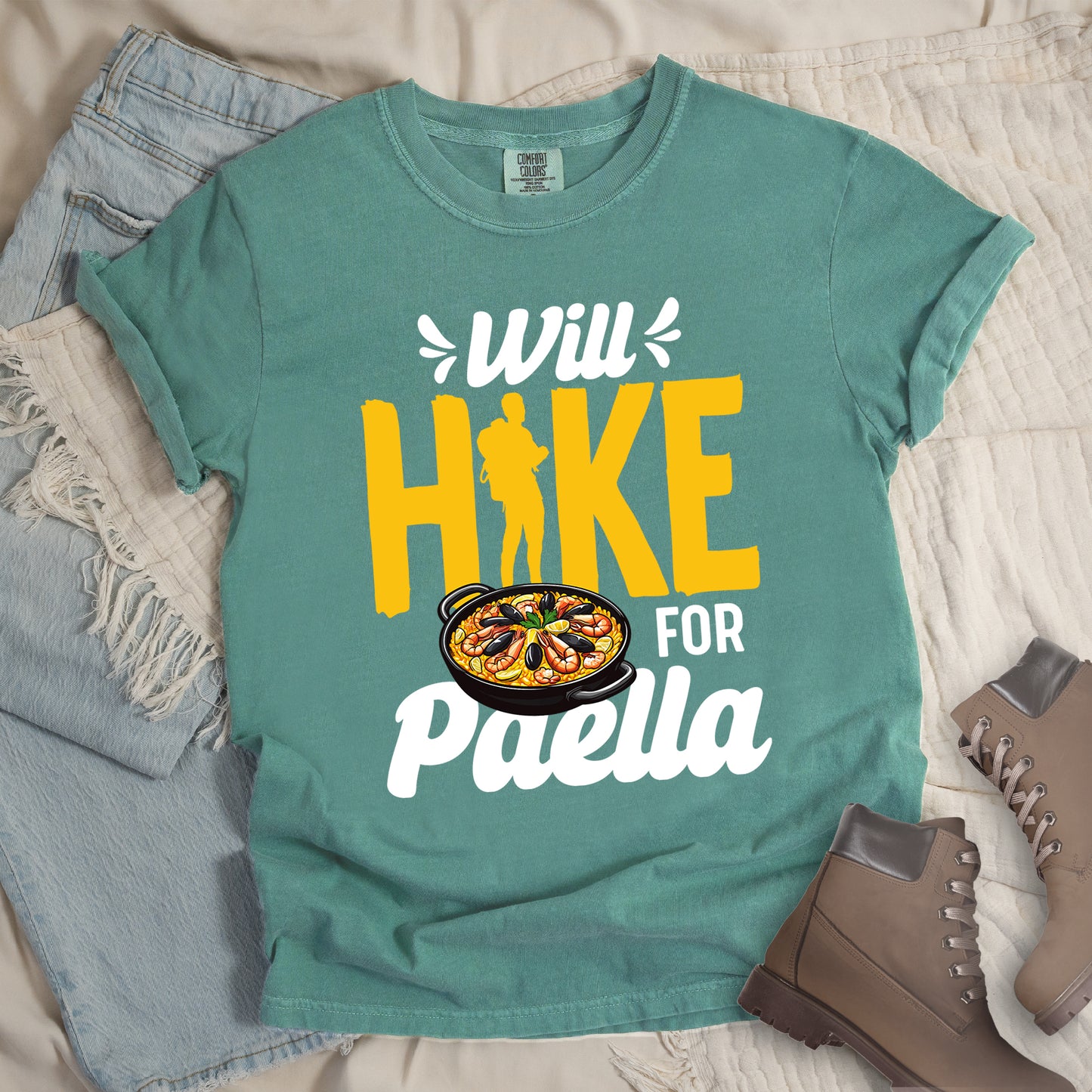 Light Green t-shirt with white and yellow text reading "Will HIKE FOR Paella". "Will" appears in white script at the top with small decorative accents, "HIKE" in large yellow letters with a yellow hiker silhouette integrated into the letter "I", "FOR" in white, and "Paella" in white script at the bottom. Features an illustration of a paella dish with seafood in a black pan to the left of "FOR" and above "Paella".