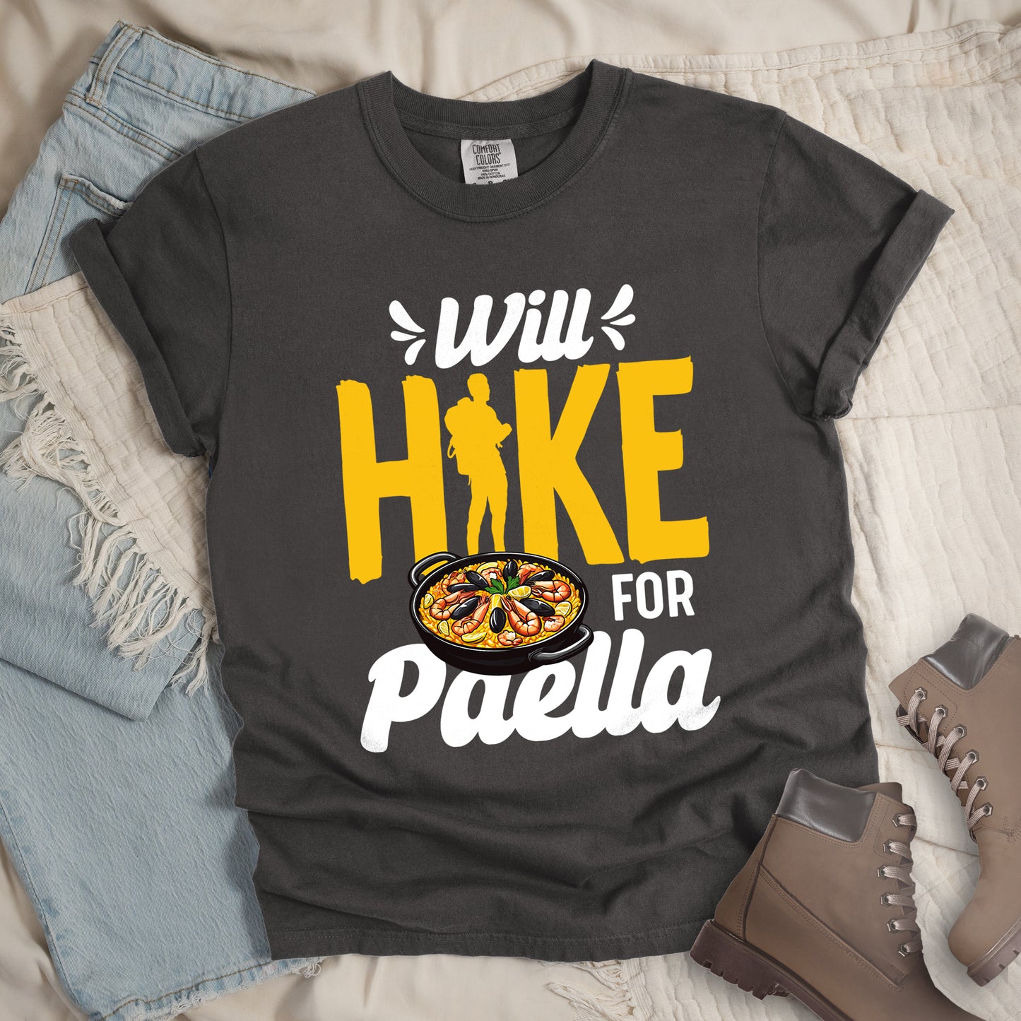 Pepper (Dark Gray) t-shirt with white and yellow text reading "Will HIKE FOR Paella". "Will" appears in white script at the top with small decorative accents, "HIKE" in large yellow letters with a yellow hiker silhouette integrated into the letter "I", "FOR" in white, and "Paella" in white script at the bottom. Features an illustration of a paella dish with seafood in a black pan to the left of "FOR" and above "Paella".