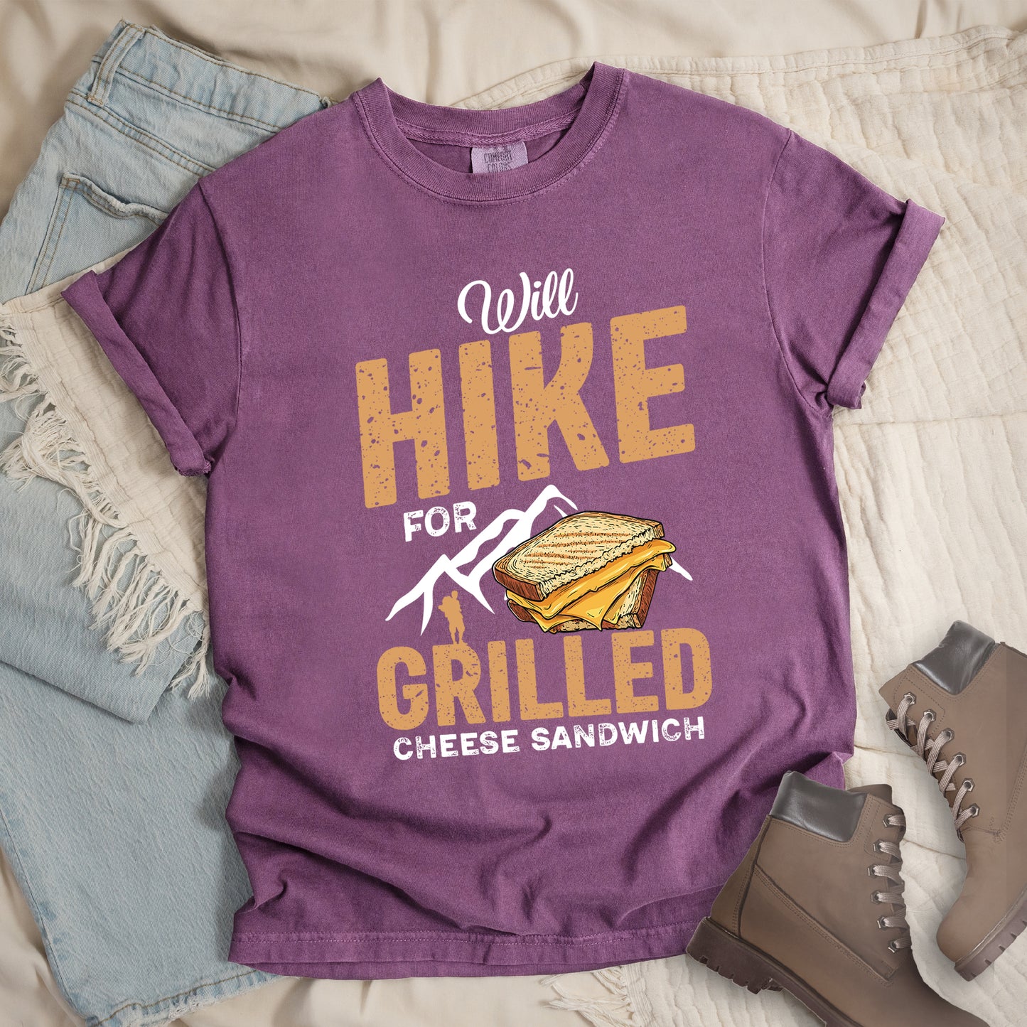 Berry (Dark Violet) t-shirt with white and golden-brown distressed text reading "Will HIKE FOR GRILLED CHEESE SANDWICH". "Will" appears in white script at the top, "HIKE" in large bold distressed golden-brown block letters, "FOR" in white, "GRILLED" in bold distressed golden-brown block letters, and "CHEESE SANDWICH" in white at the bottom. Features white mountain silhouettes, a golden-brown hiker silhouette, and an illustration of a grilled cheese sandwich with toasted bread and yellow cheese inside.