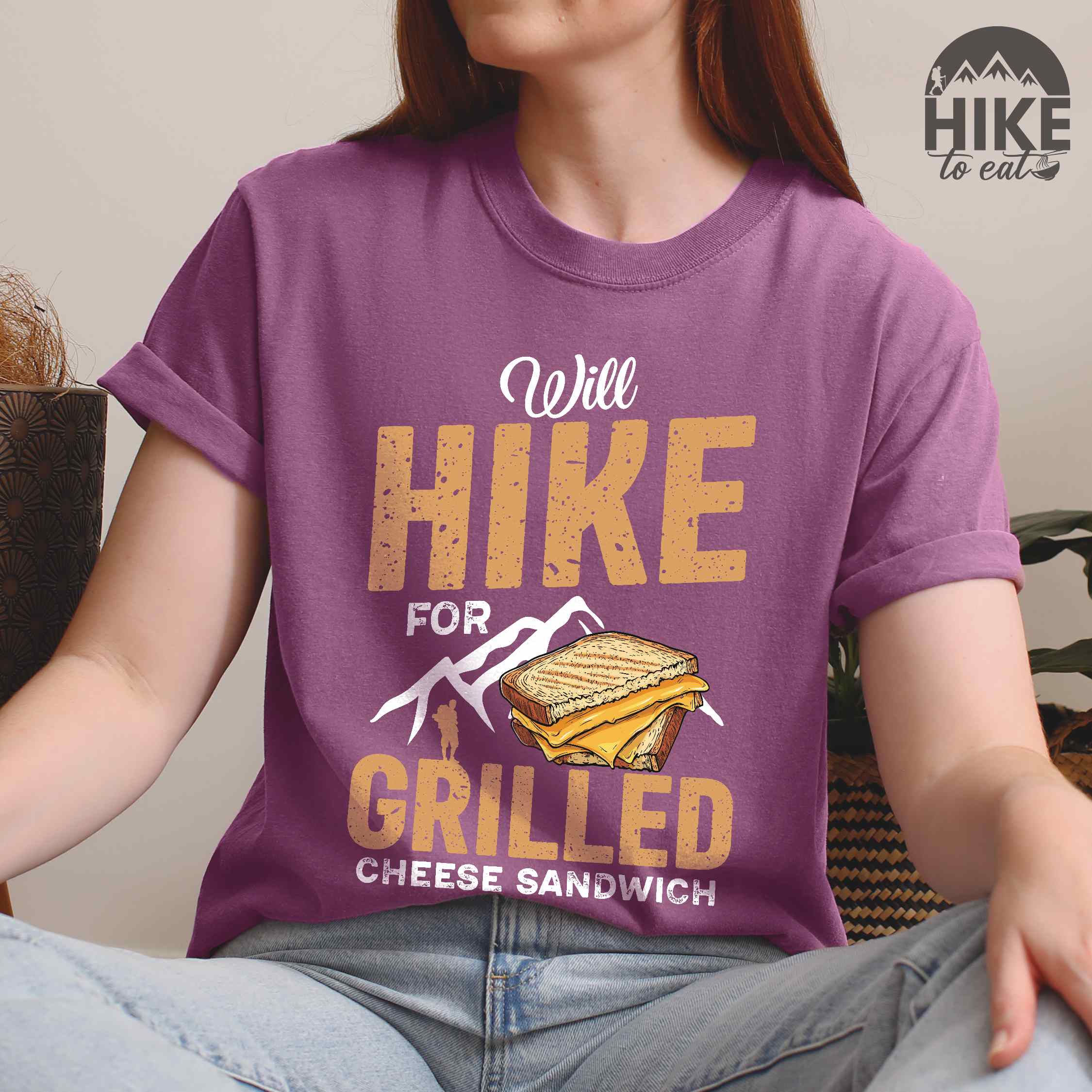 Woman wearing berry "will hike for grilled cheese sandwich" colored vintage tee with bold typography, mountains, birds, illustrated grilled cheese sandwich, and backpackers for sandwich enthusiasts
