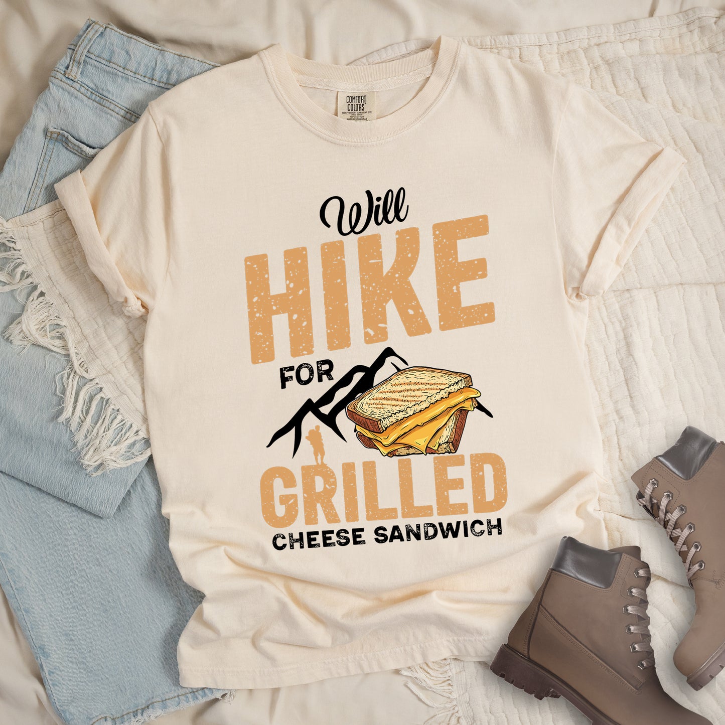 Ivory (Cream White) t-shirt with black and golden-brown distressed text reading "Will HIKE FOR GRILLED CHEESE SANDWICH". "Will" appears in black script at the top, "HIKE" in large bold distressed golden-brown block letters, "FOR" in black, "GRILLED" in bold distressed golden-brown block letters, and "CHEESE SANDWICH" in black at the bottom. Features black mountain silhouettes, a golden-brown hiker silhouette, and an illustration of a grilled cheese sandwich with toasted bread and yellow cheese inside.