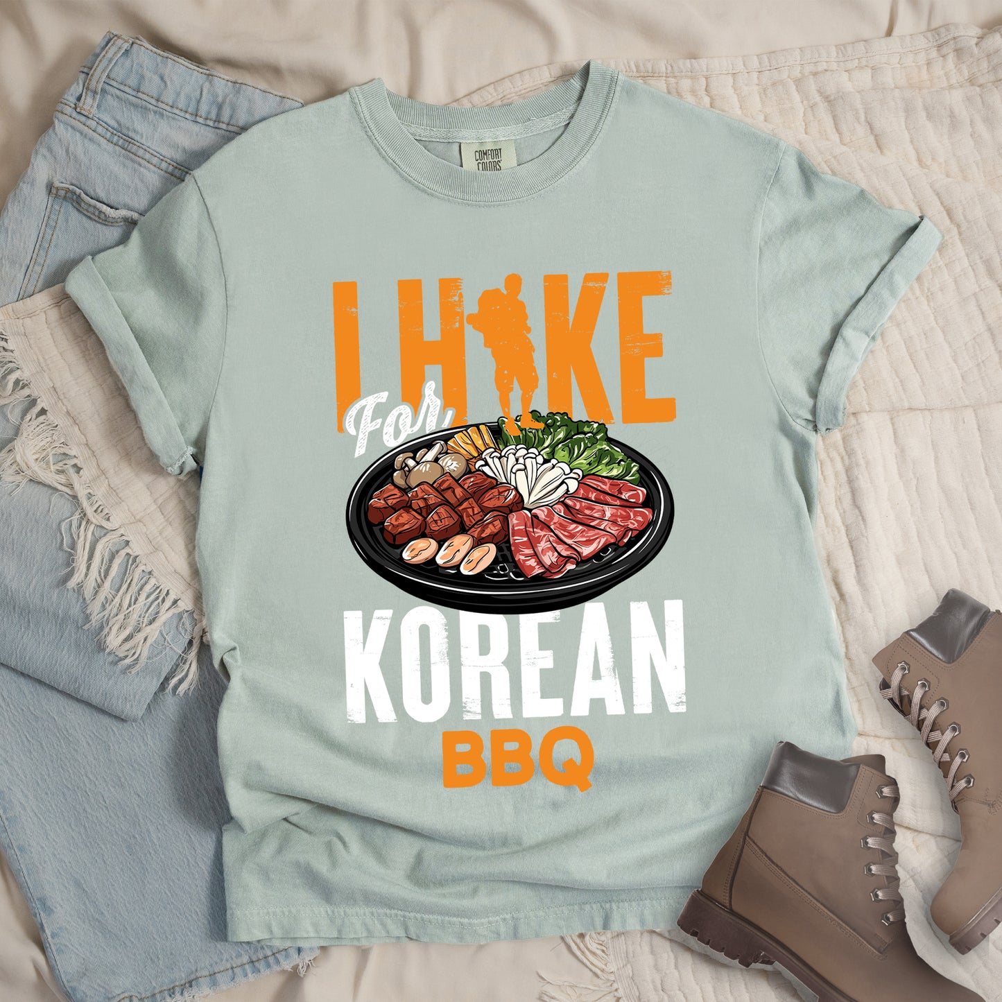 Bay (Light Gray) t-shirt with orange and white text reading "I HIKE For KOREAN BBQ". "I HIKE" appears in large distressed orange letters at the top with a hiker silhouette integrated into the letter "I", "For" in white script in the middle, "KOREAN" in large distressed white letters, and "BBQ" in orange at the bottom. Features a detailed illustration of Korean barbecue with meat, mushrooms, and vegetables on a black plate in between "For" and "KOREAN."