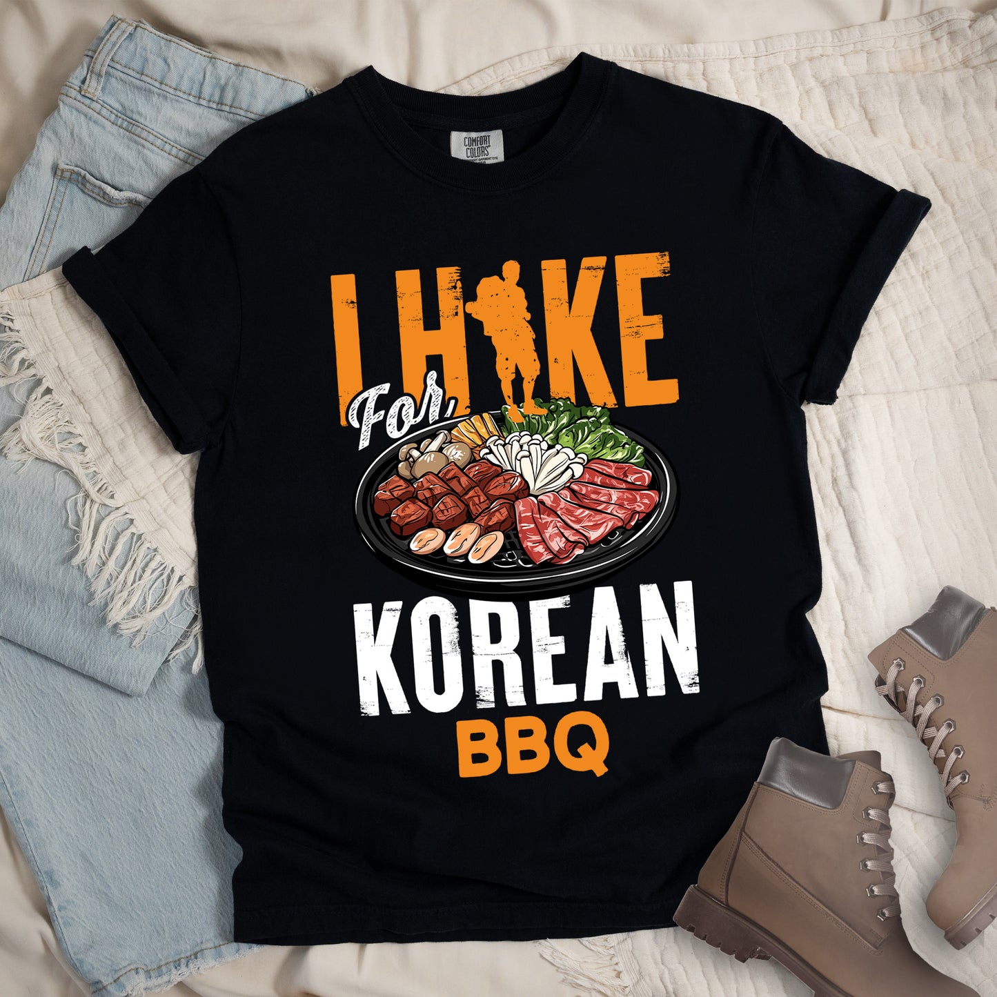 Black t-shirt with orange and white text reading "I HIKE For KOREAN BBQ". "I HIKE" appears in large distressed orange letters at the top with a hiker silhouette integrated into the letter "I", "For" in white script in the middle, "KOREAN" in large distressed white letters, and "BBQ" in orange at the bottom. Features a detailed illustration of Korean barbecue with meat, mushrooms, and vegetables on a black plate in between "For" and "KOREAN."