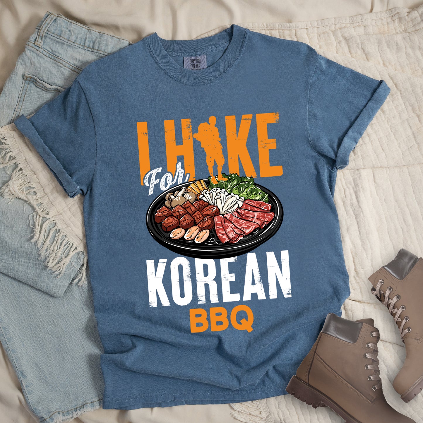 Blue Jean (Denim Blue) t-shirt with orange and white text reading "I HIKE For KOREAN BBQ". "I HIKE" appears in large distressed orange letters at the top with a hiker silhouette integrated into the letter "I", "For" in white script in the middle, "KOREAN" in large distressed white letters, and "BBQ" in orange at the bottom. Features a detailed illustration of Korean barbecue with meat, mushrooms, and vegetables on a black plate in between "For" and "KOREAN."