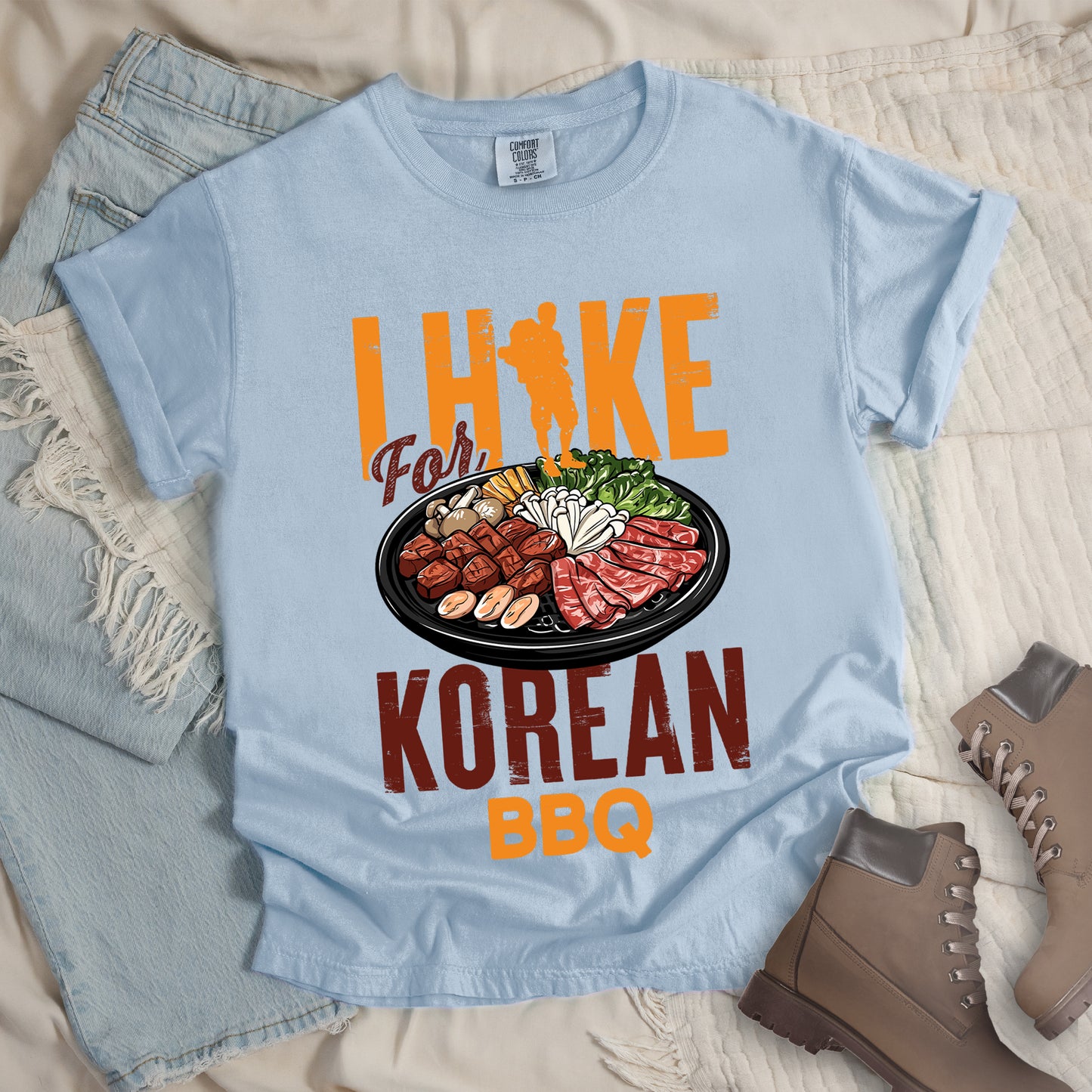 Chambray (Light Blue) t-shirt with orange and red text reading "I HIKE For KOREAN BBQ". "I HIKE" appears in large distressed orange letters at the top with a hiker silhouette integrated into the letter "I", "For" in red script in the middle, "KOREAN" in large distressed red letters, and "BBQ" in orange at the bottom. Features a detailed illustration of Korean barbecue with meat, mushrooms, and vegetables on a black plate in between "For" and "KOREAN."