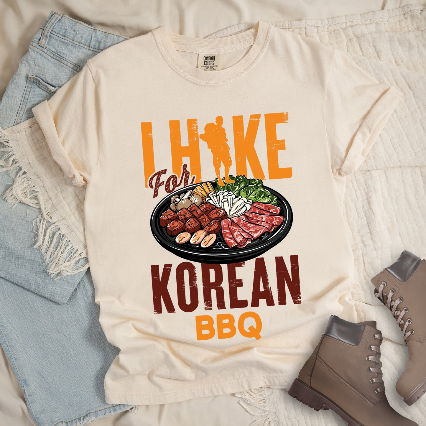 Ivory (Cream White) t-shirt with orange and red text reading "I HIKE For KOREAN BBQ". "I HIKE" appears in large distressed orange letters at the top with a hiker silhouette integrated into the letter "I", "For" in red script in the middle, "KOREAN" in large distressed red letters, and "BBQ" in orange at the bottom. Features a detailed illustration of Korean barbecue with meat, mushrooms, and vegetables on a black plate in between "For" and "KOREAN."