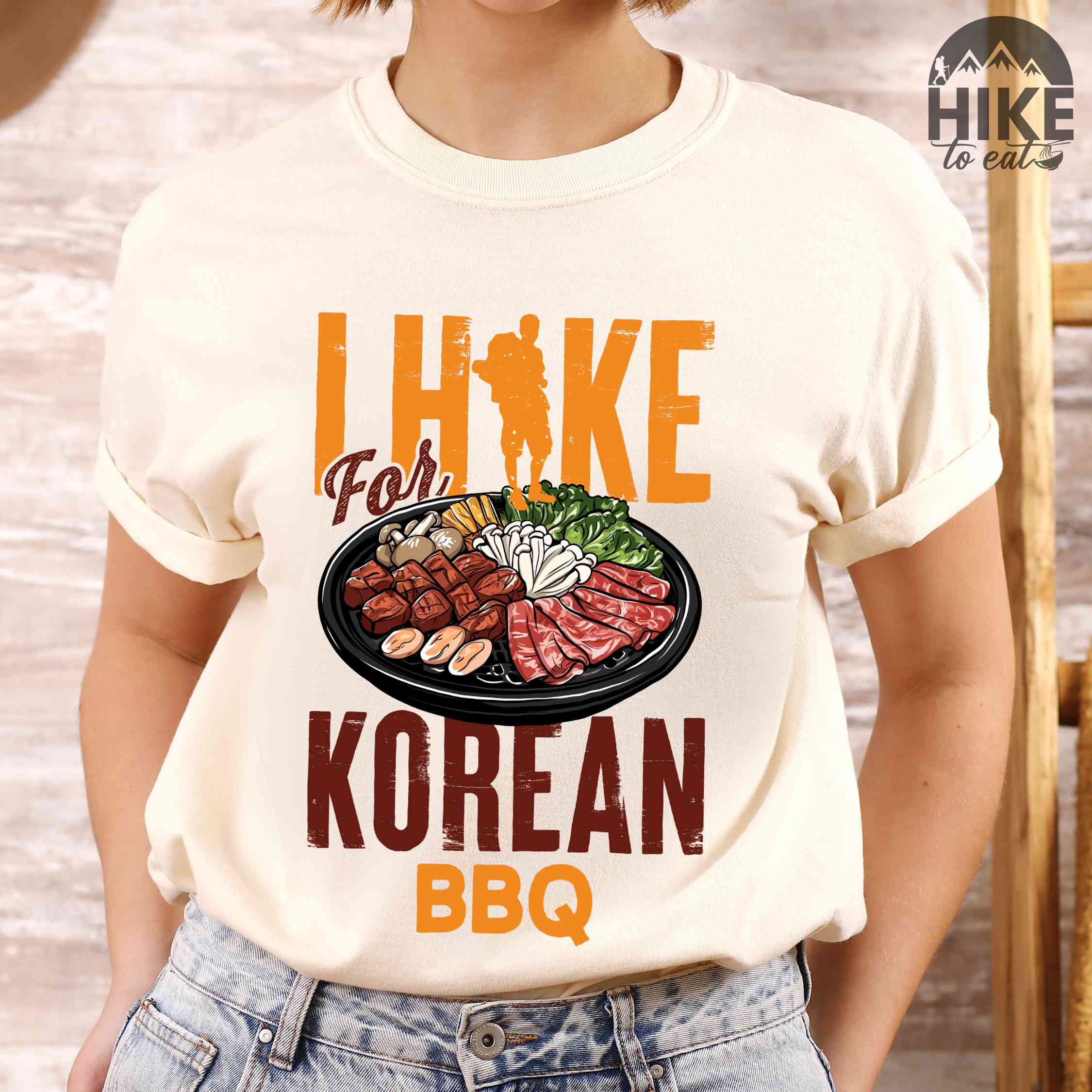 Woman wearing ivory "I hike for korean bbq" colored vintage tee with bold typography, illustrated grilling platter korean bbq, mountains, birds, and backpacker for kbbq lovers.