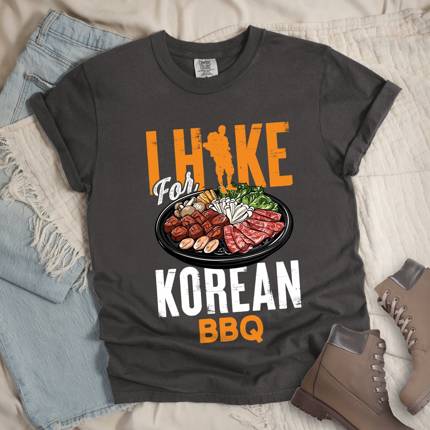 Pepper (Dark Gray) t-shirt with orange and white text reading "I HIKE For KOREAN BBQ". "I HIKE" appears in large distressed orange letters at the top with a hiker silhouette integrated into the letter "I", "For" in white script in the middle, "KOREAN" in large distressed white letters, and "BBQ" in orange at the bottom. Features a detailed illustration of Korean barbecue with meat, mushrooms, and vegetables on a black plate in between "For" and "KOREAN."