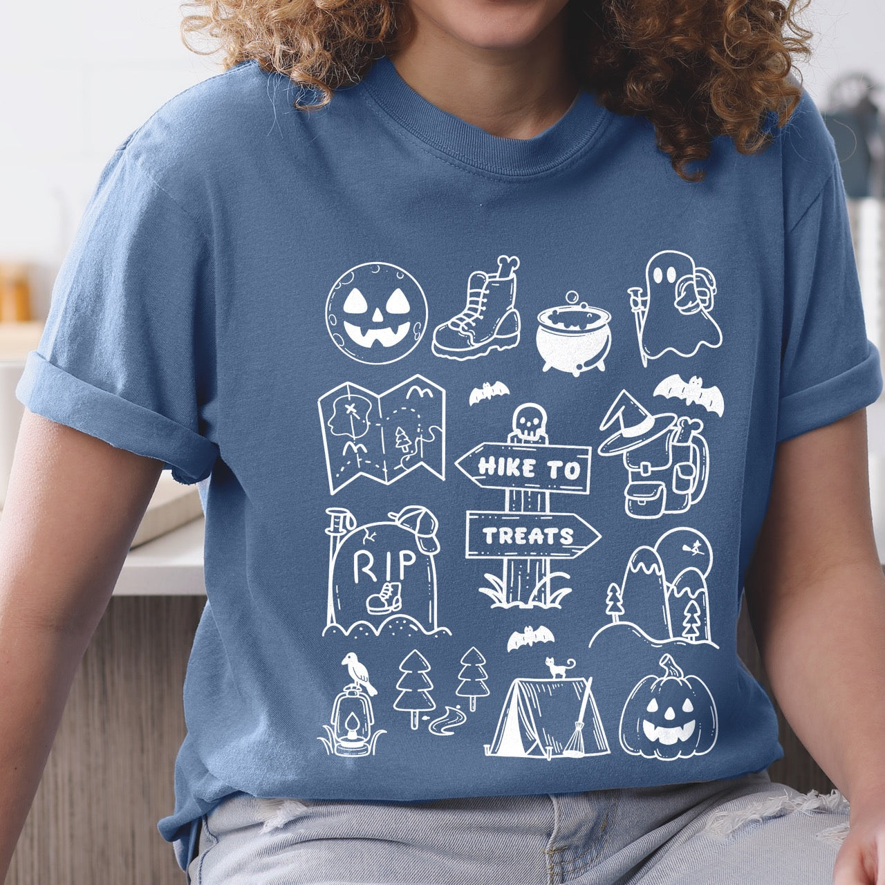 woman wearing Blue Jean (blue) Halloween adventure vintage tee featuring illustrated icon collection with jack-o-lantern boots cauldron ghost backpacker map RIP tombstone Hike to Treats trail sign tent pumpkin bats and trees on Comfort Colors shirt for spooky season celebrations for hikers, foodies and adventurers