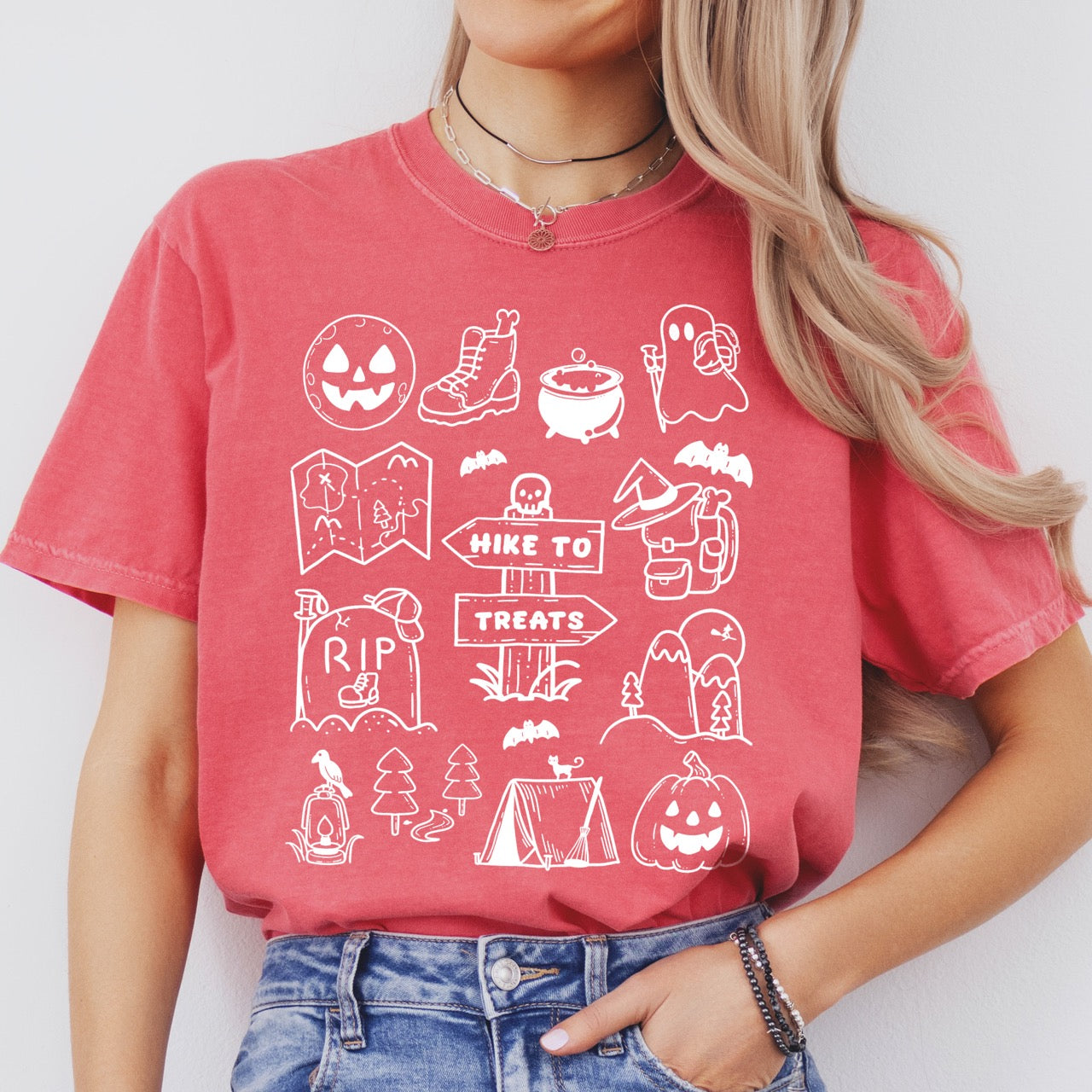 Woman wearing Watermelon (coral) Halloween adventure vintage tee featuring illustrated icon collection with jack-o-lantern boots cauldron ghost backpacker map RIP tombstone Hike to Treats trail sign tent pumpkin bats and trees on Comfort Colors shirt for spooky season celebrations for hikers, foodies and adventurers