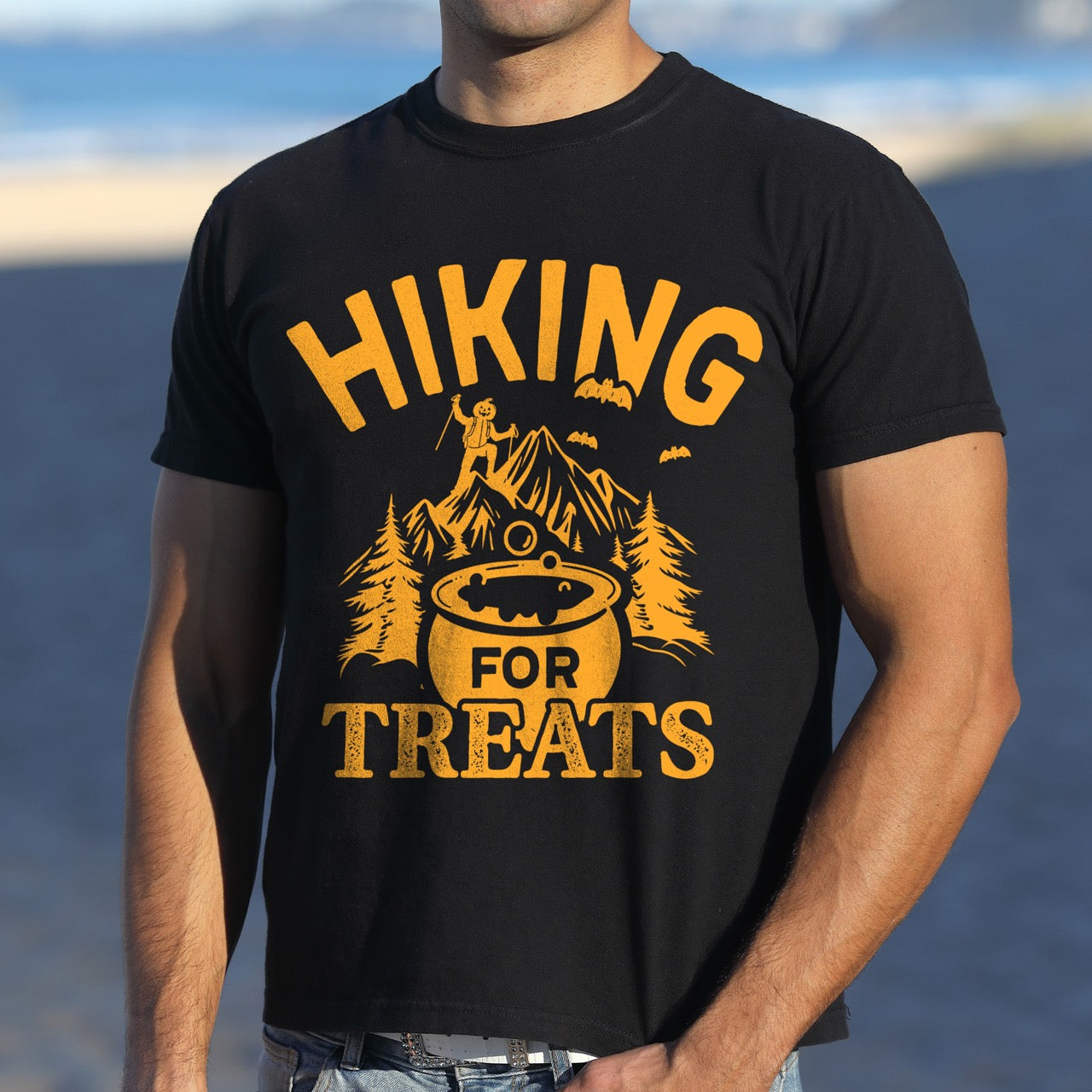 Man wearing Black Halloween vintage tee Hiking for Treats featuring curved typography bubbling cauldron with candy mountain landscape with character on peak pine trees and flying bats on Comfort Colors shirt for spooky season celebrations for hikers, foodies and adventurers.